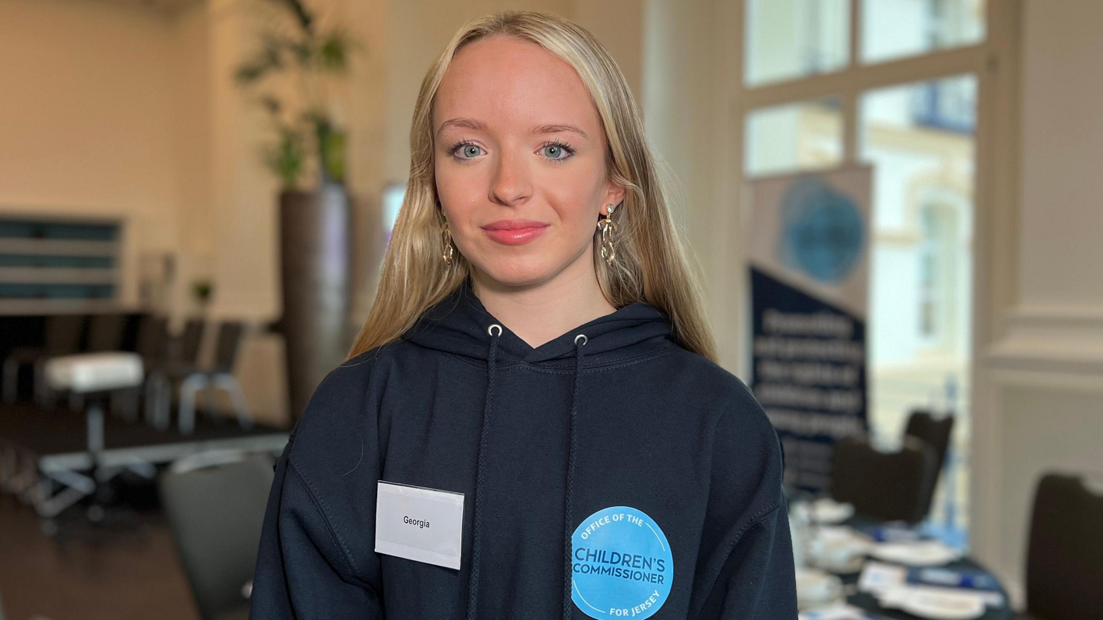 Georgia looks at the camera wearing a dark blue hoodie and behind her is a room with tables and chairs event up for the event. Her blonde hair comes below her shoulders.