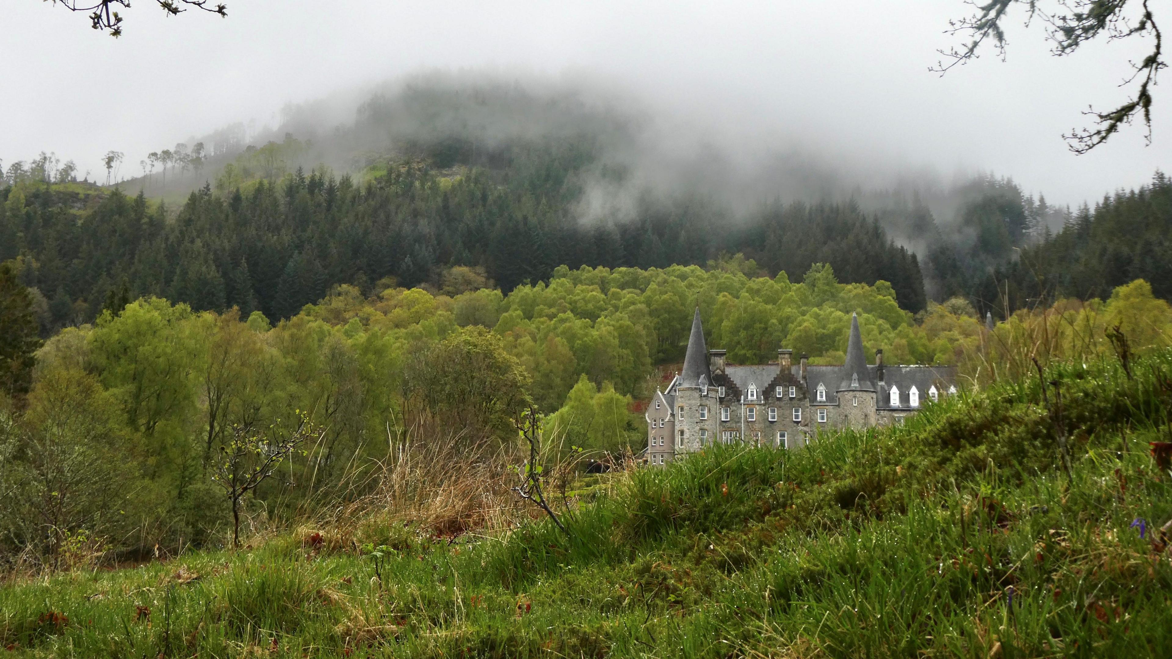Huge grey house with two pointy roofs nestled in green countrside beside a hill covered in trees.