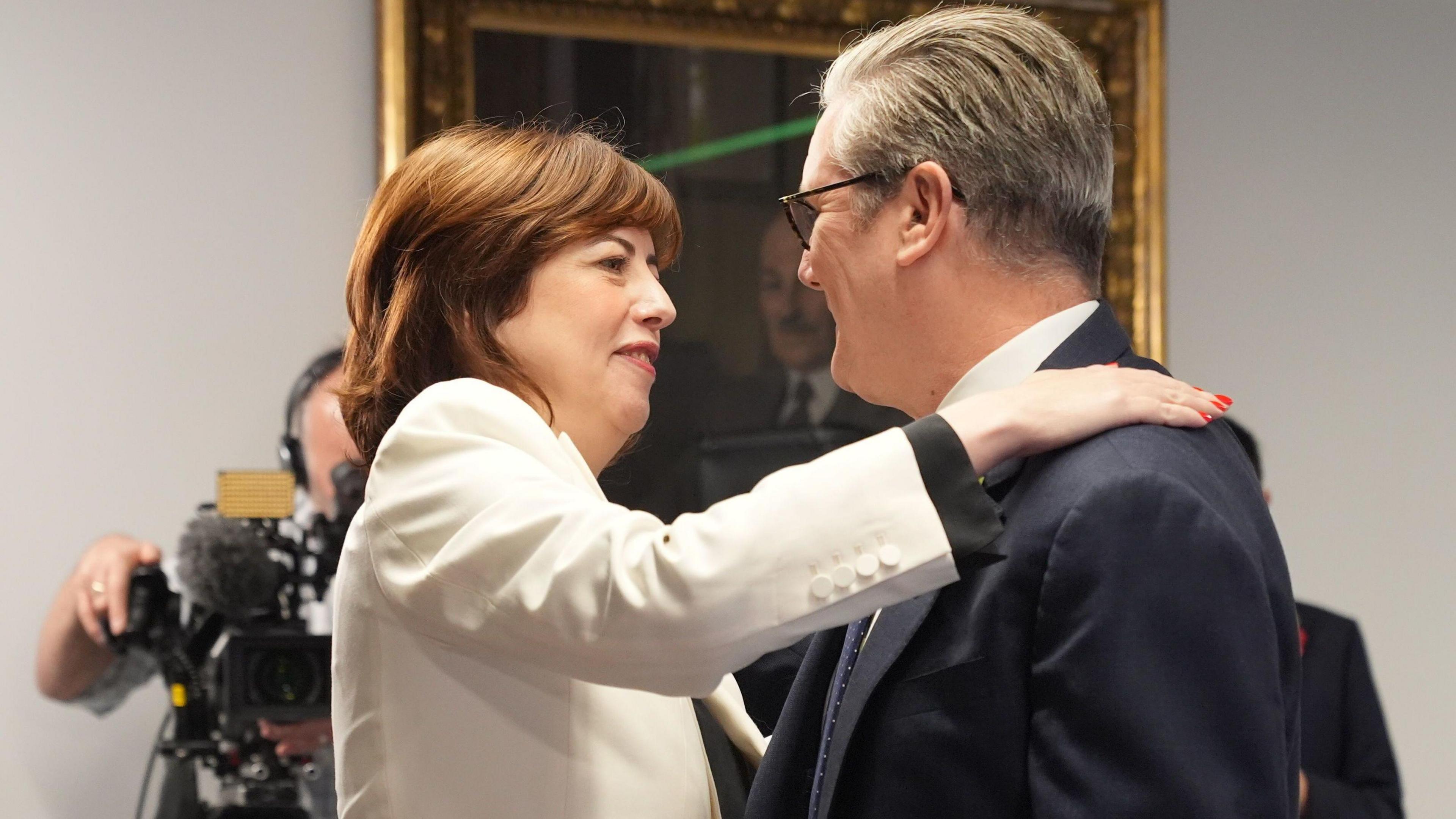 Lucy Powell, new deputy Labour leader, embracing Prime Minister Sir Keir Starmer indoors. Lucy Powell is wearing a light-colored blazer, and Sir Keir Starmer is dressed in a dark suit.