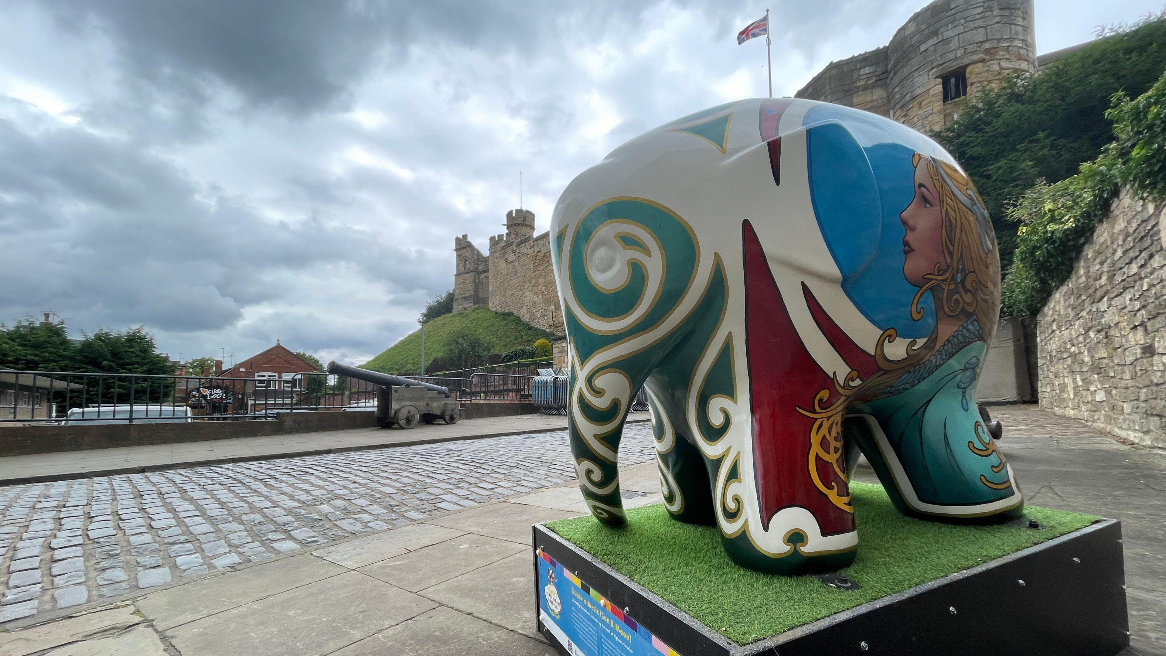 A statue of Elmer the elephant standing on a plinth outside Lincoln Castle and has the face of a woman on the back and a green swirling effect around his eyes and trunk