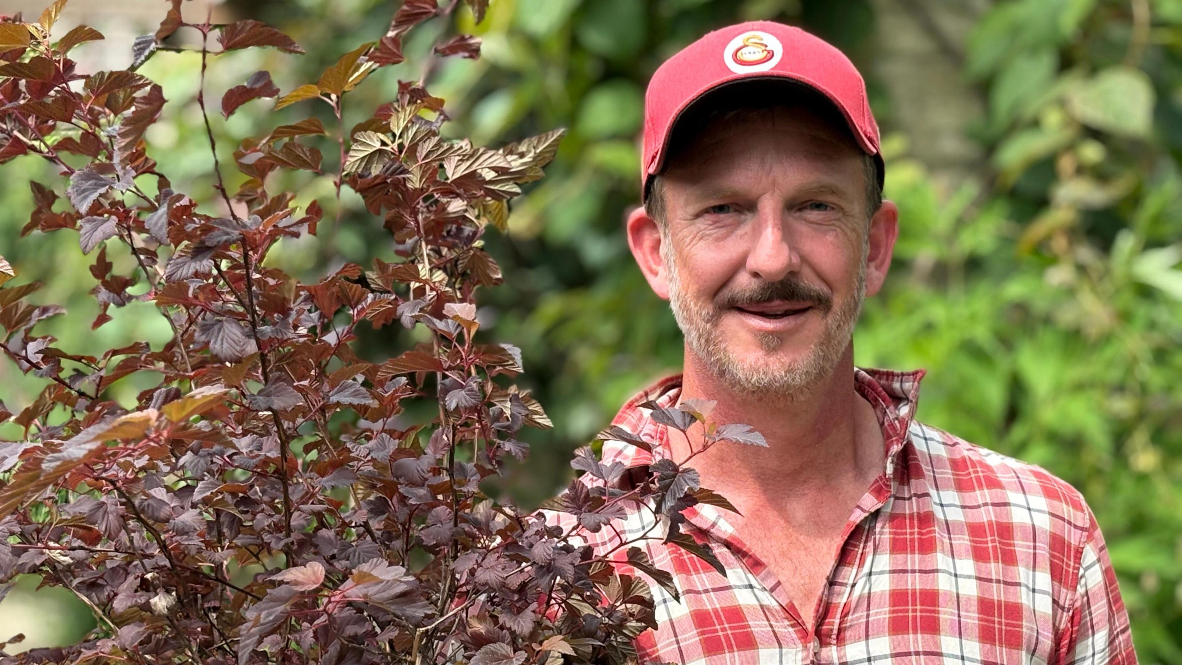 A man in a red and white checked shirt and red baseball cap holds a purple plant.