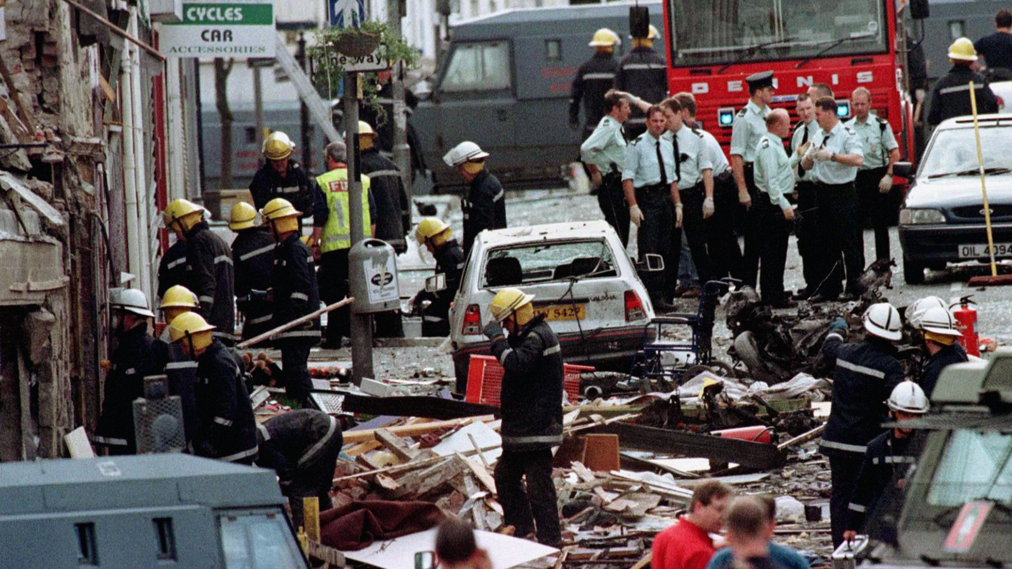 The picture shows the aftermath of the Omagh bomb. Firefighters and police officers work through the wreckage in the town with parts of buildings lying on the ground and a car with its windows blown out