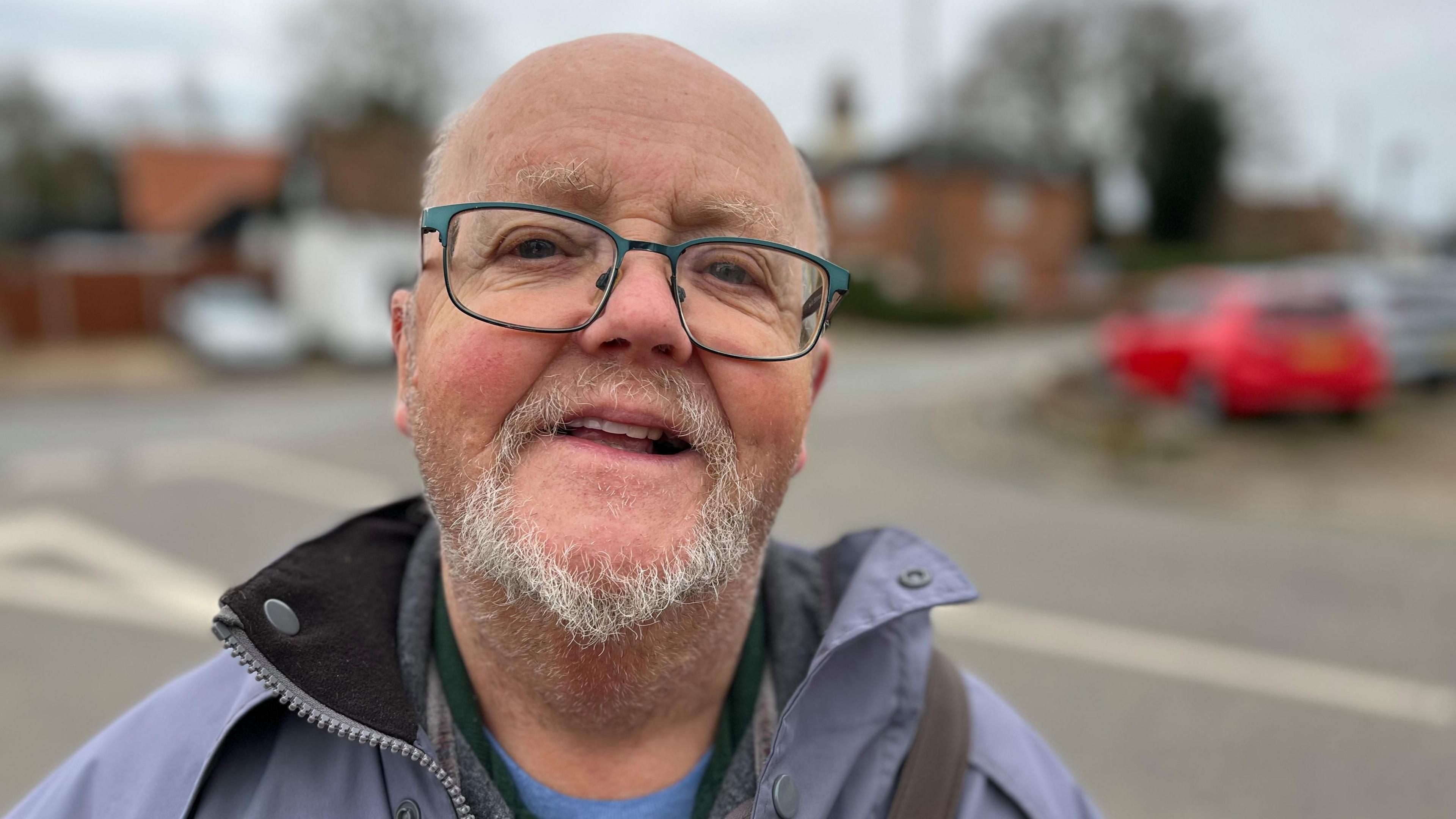 Peter Robertson has short, white hair and a white goatee beard. He is wearing glasses and is looking at the camera.
He is wearing a lilac jacket with a blue t shirt underneath.