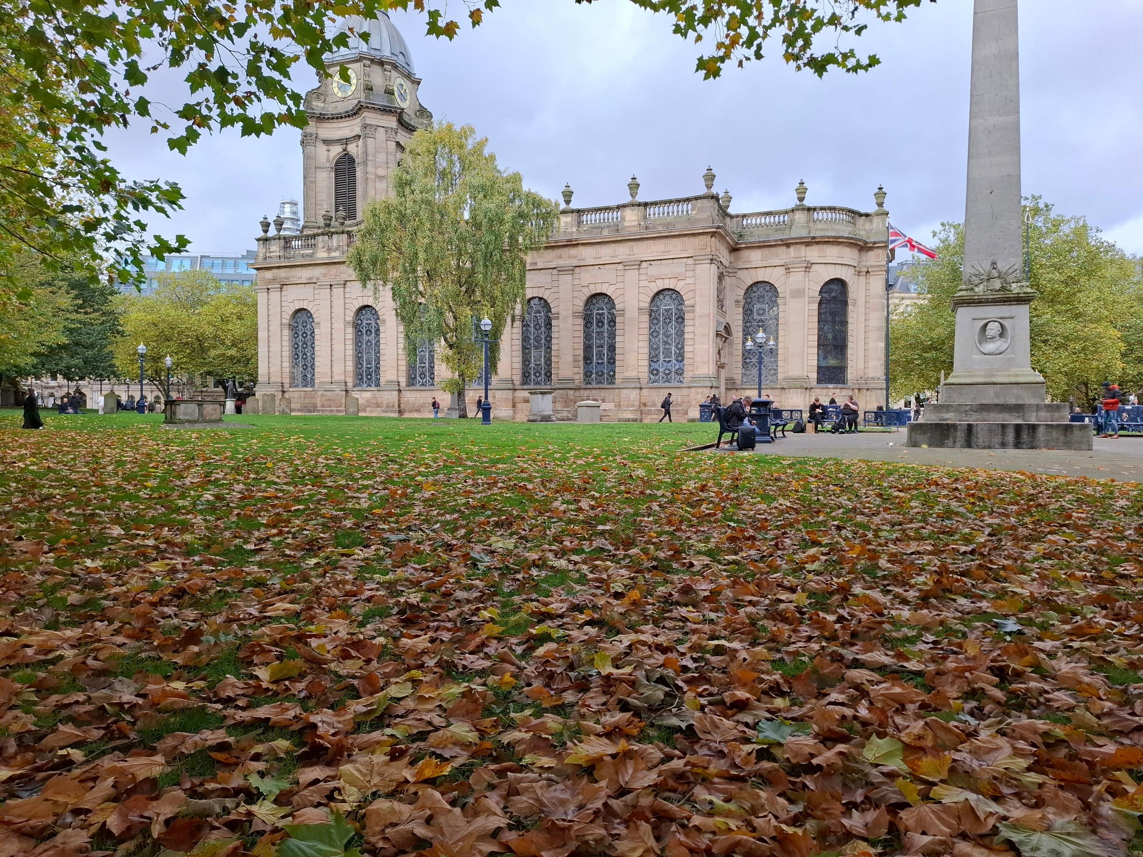 Brown leaves scattered on grass, surrounding a church with a domed spire. To one side, an obelisk stands in the grounds, and a union jack is flown.