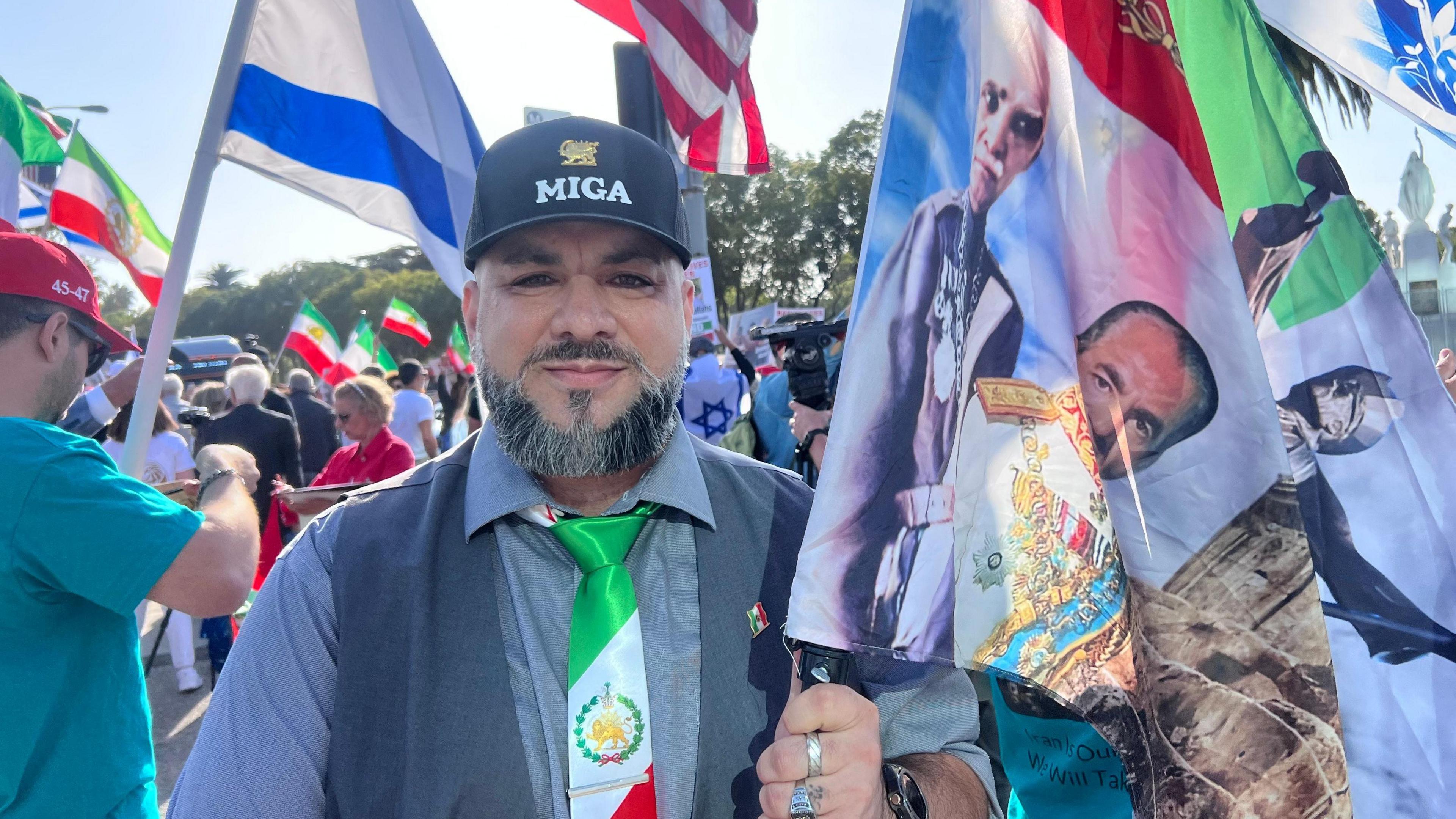 A man dressed in a MIGA – Make Iran Great Again – hat with a tie showing the lion and sun emblem from Iran's pre-1979 flag - poses carrying several flags at a protest in West LA. You can see a crowd behind him and people waving US, Iranian and Israeli flags.