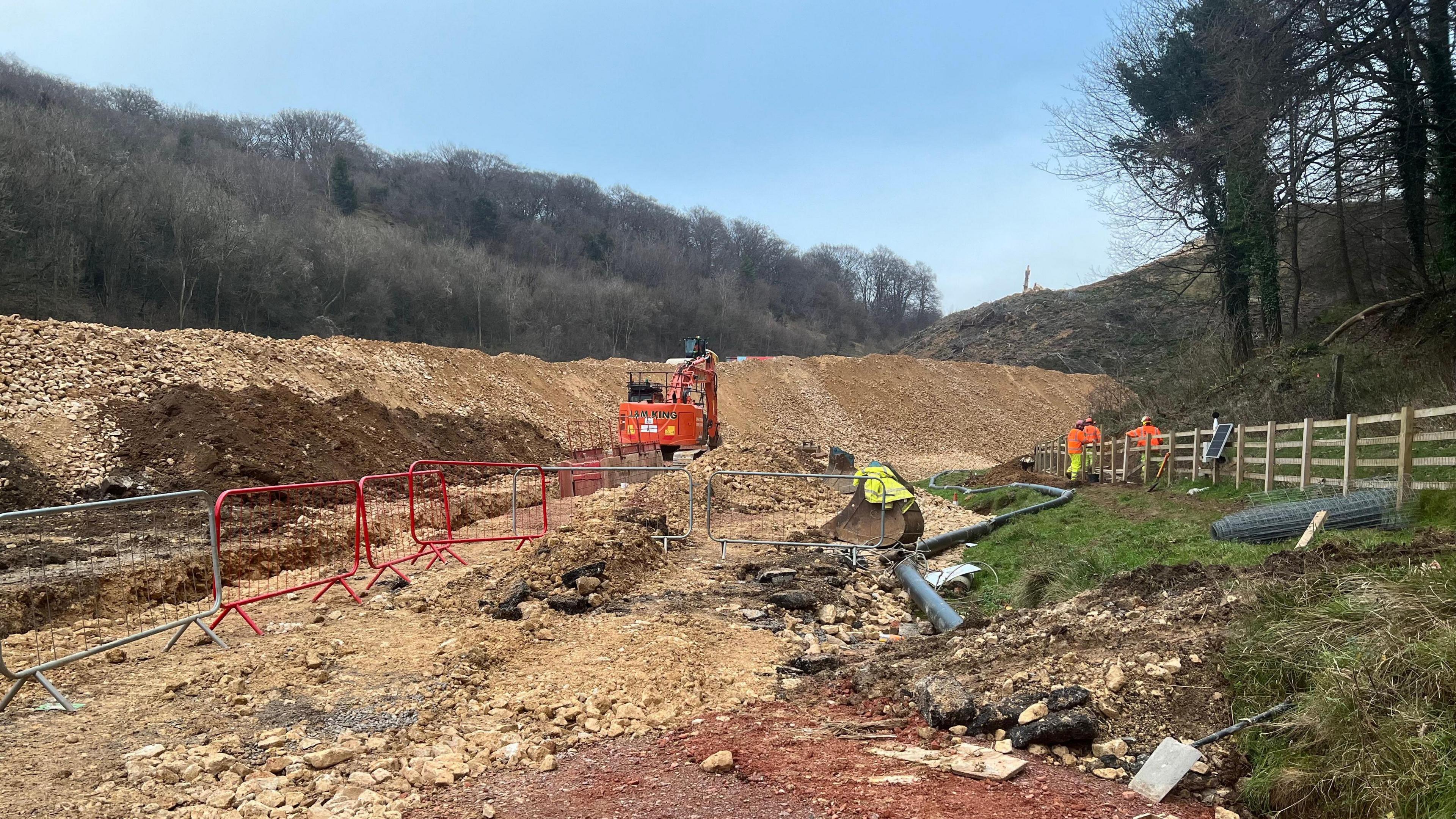 A construction site in Gloucestershire for the Missing Link scheme. Construction vehicles and machines can be seen, as well as debris. A few workers can be seen to the side of the frame and they are all dressed in high vis clothing. 