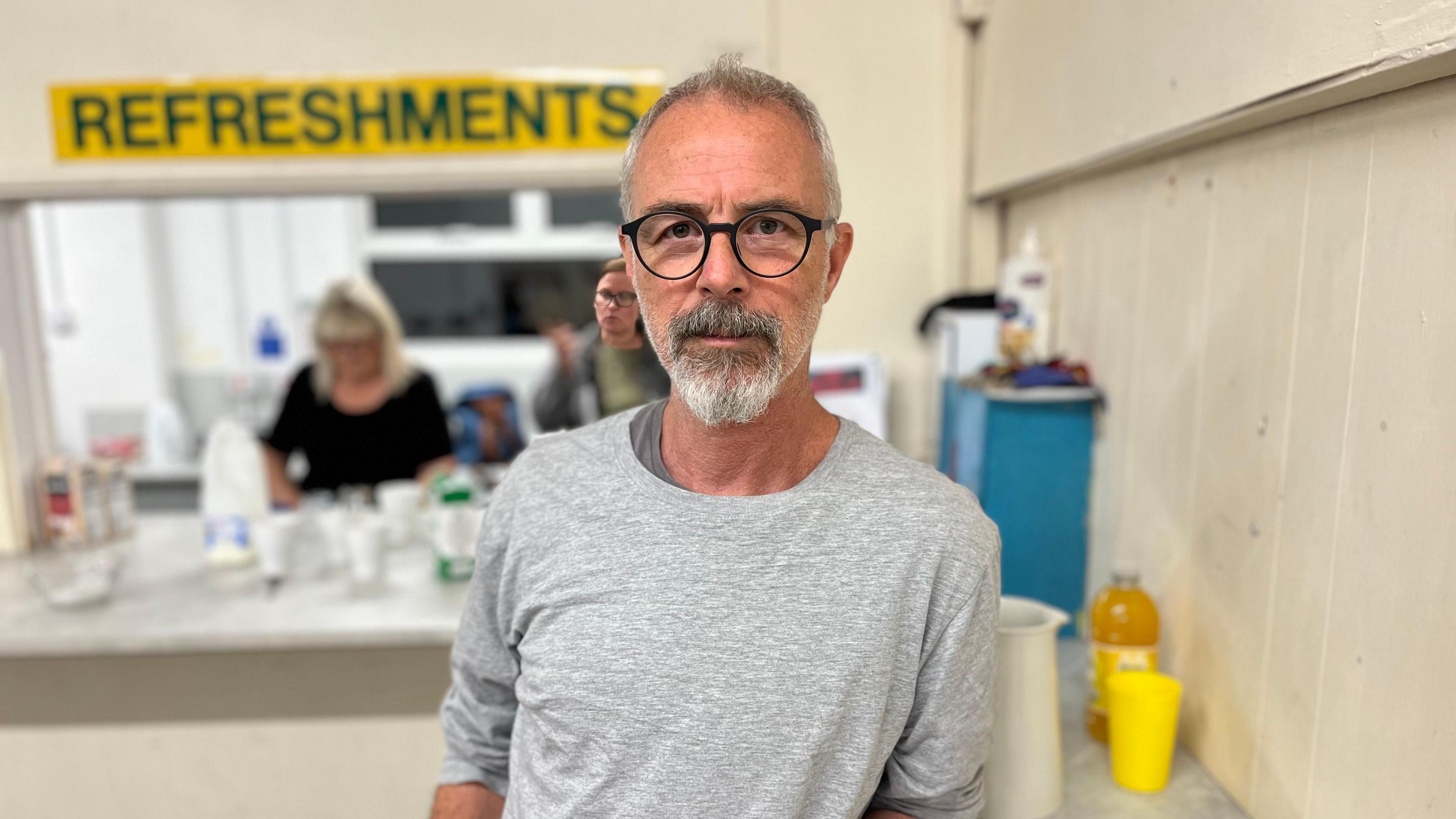 Peter, who has grey hair and a grey beard, looks at the camera. He wears dark round glasses and a grey top. Behind him is a refreshments hatch inside the community centre.