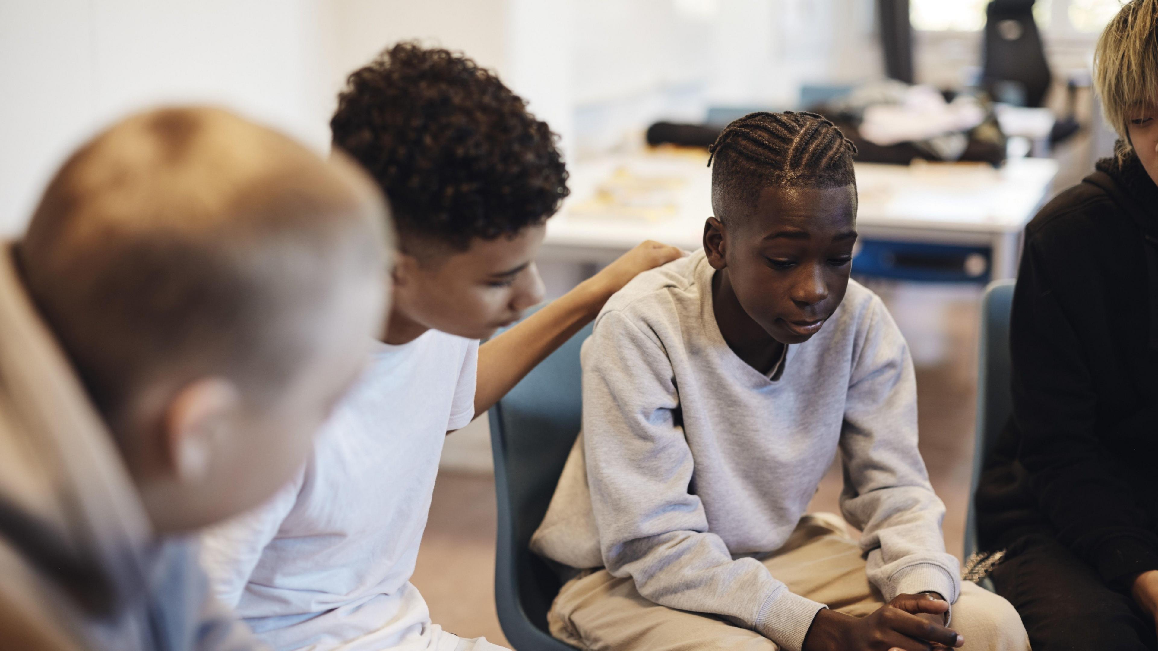A young boy is sat down looking pensive. He is wearing a grey jumper and pale brown trousers. A boy sat next to him, wearing a white t-shirt, has his hand on the first boy's back. There is a blurred head in the foreground and another boy just visible to the side, suggesting they are sat in a circle.