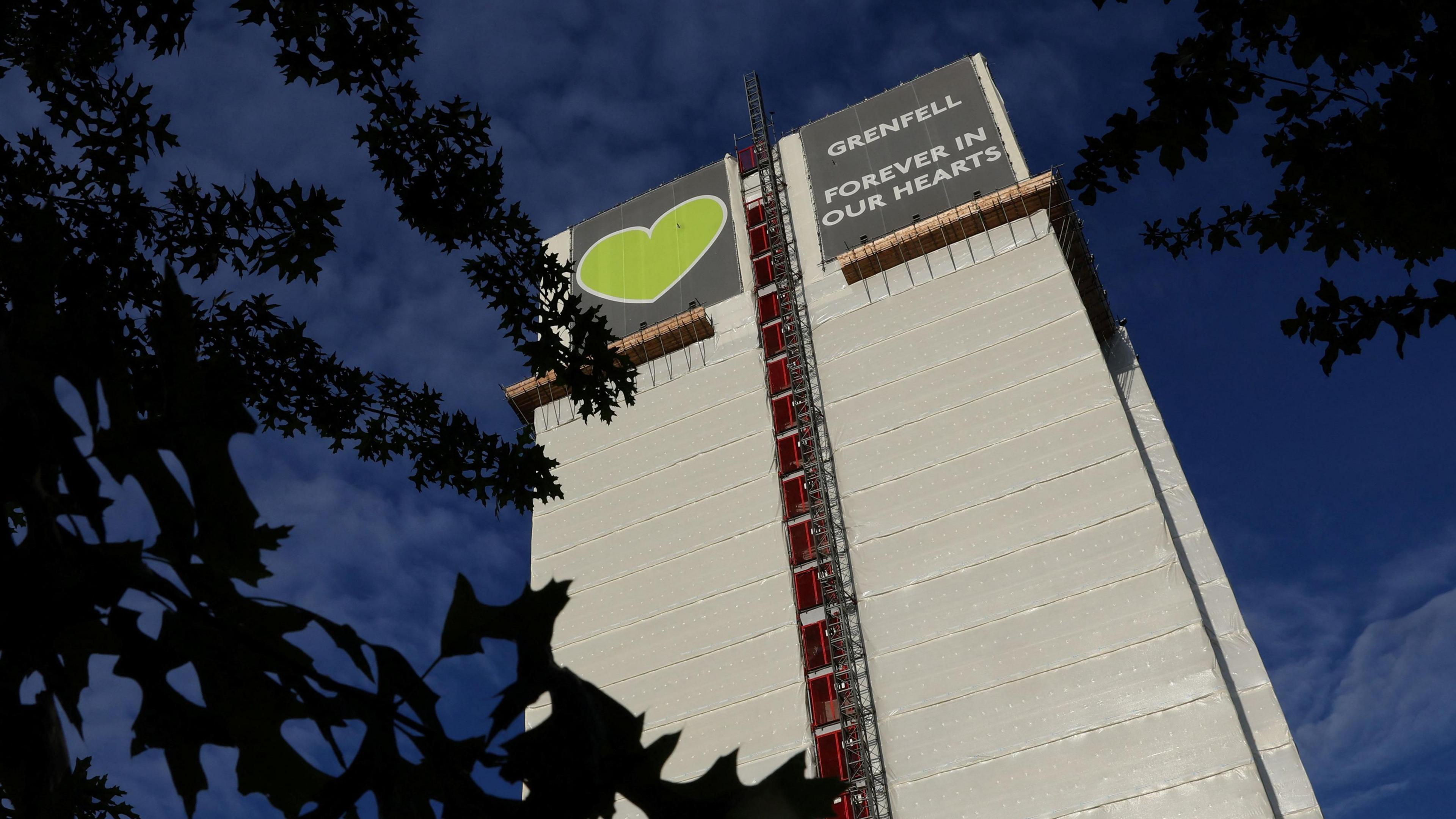 The Grenfell Tower covered in plastic sheeting. At the top is a sign with a green heart and another reading "Grenfell - forever in our hearts" in capital letters. 