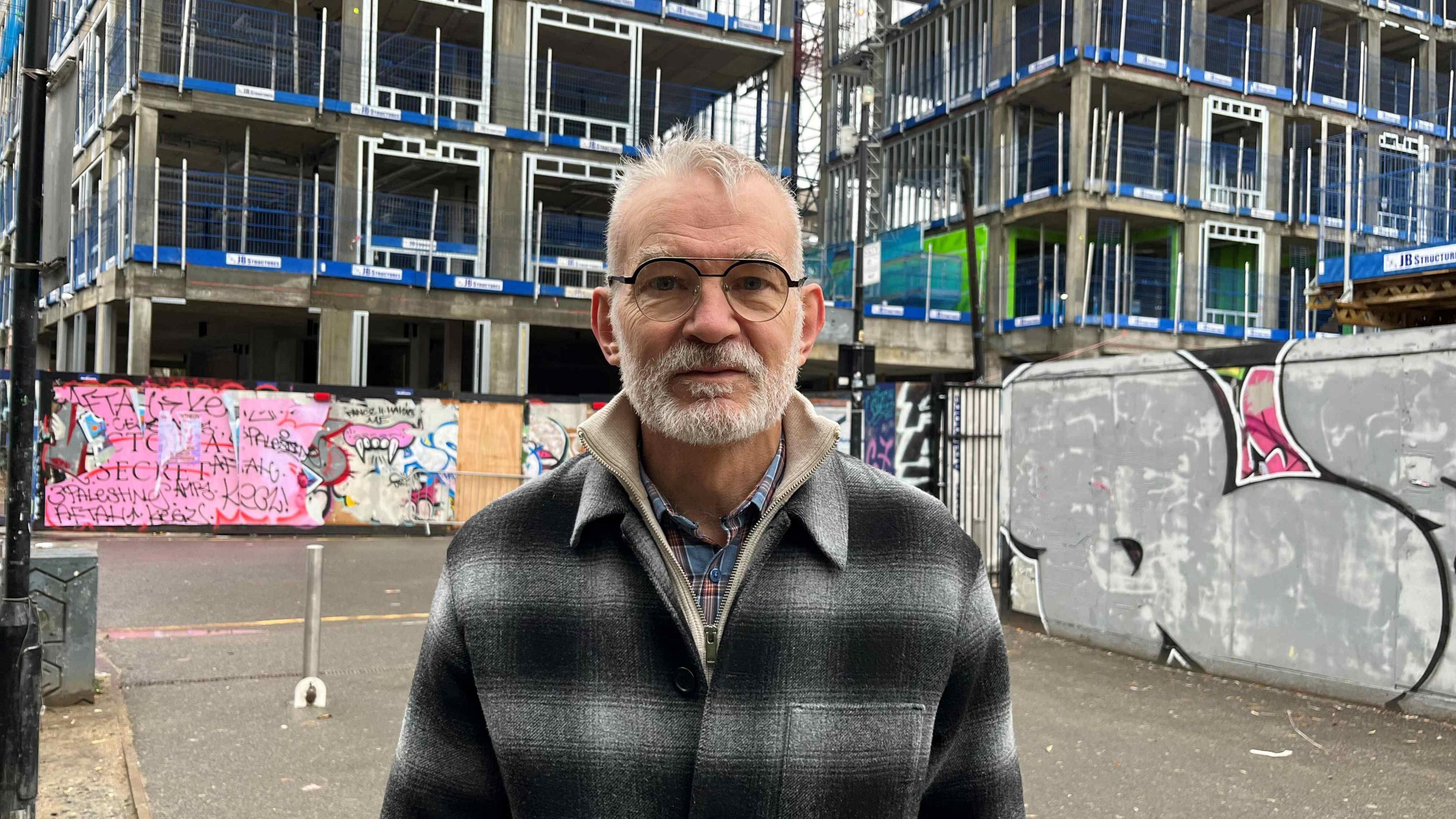 Andrew Boff, chair of the planning and regeneration committee for the London Assembly outside a construction. The block is still built so you can see the exposed framework, and at ground level are two hoardings he's standing in front or, with graffiti on them.