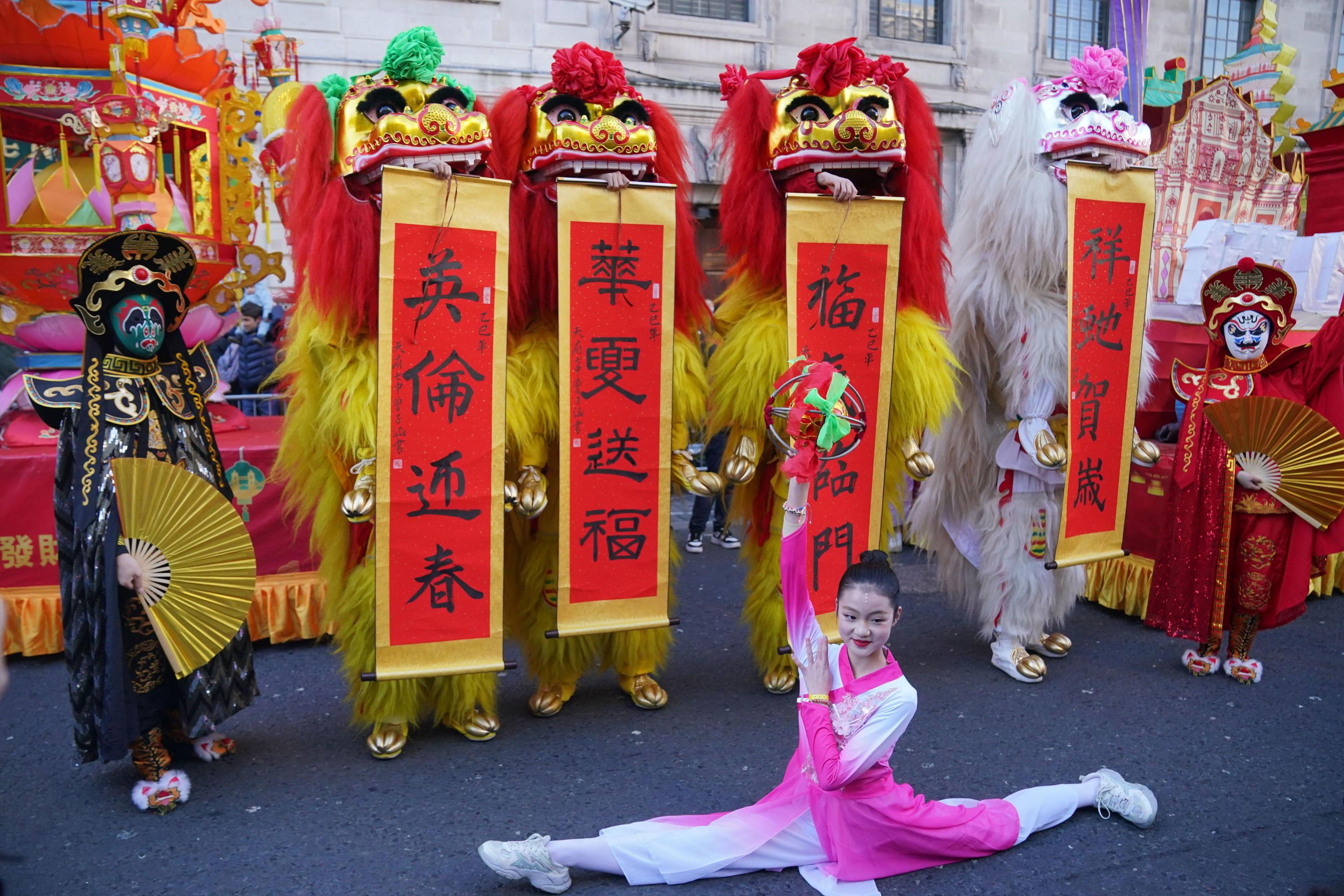 A group of lion dancers in red, yellow, and white costumes hold vertical banners with Chinese calligraphy. A young female performer in a pink and white outfit is doing the splits in front of them while holding a prop.