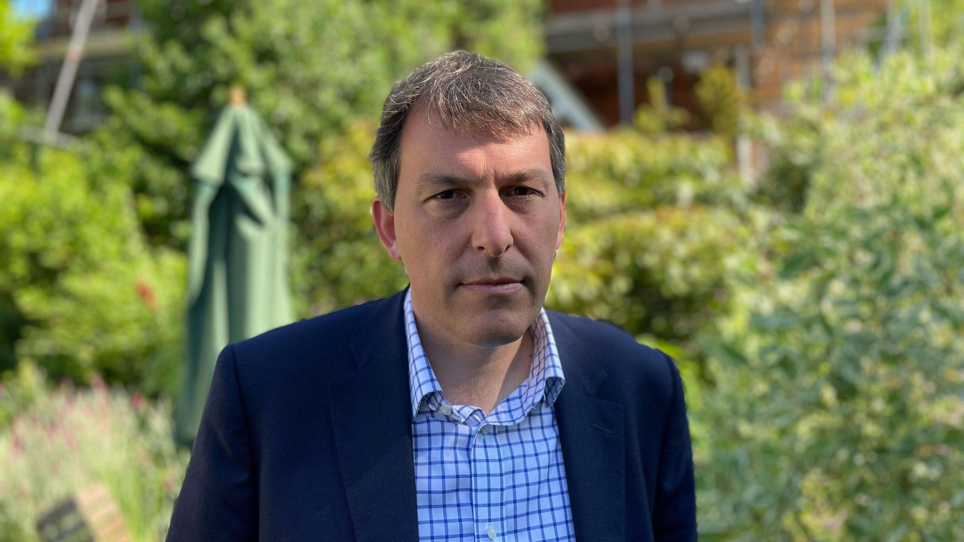 A middle-aged man stares at the camera wearing a shirt and jacket with a sun umbrella and trees in the background.