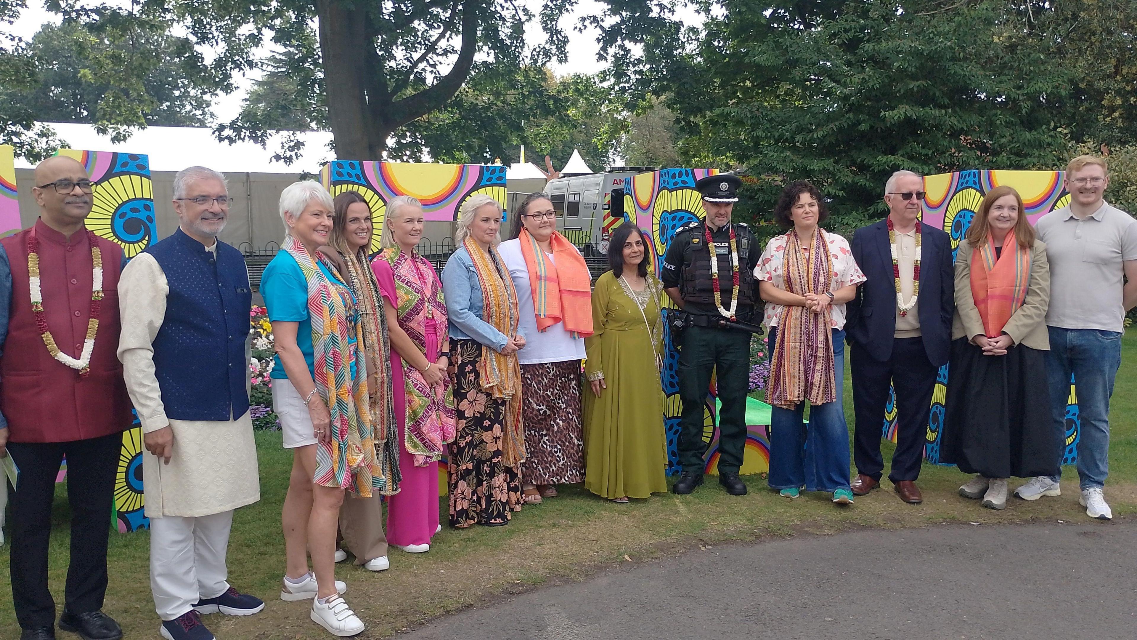 A group of people are standing together for a photo. They're in the Botanic Gardens. They're standing on grass and some trees are behind them. Some are wearing scarves or garlands.
