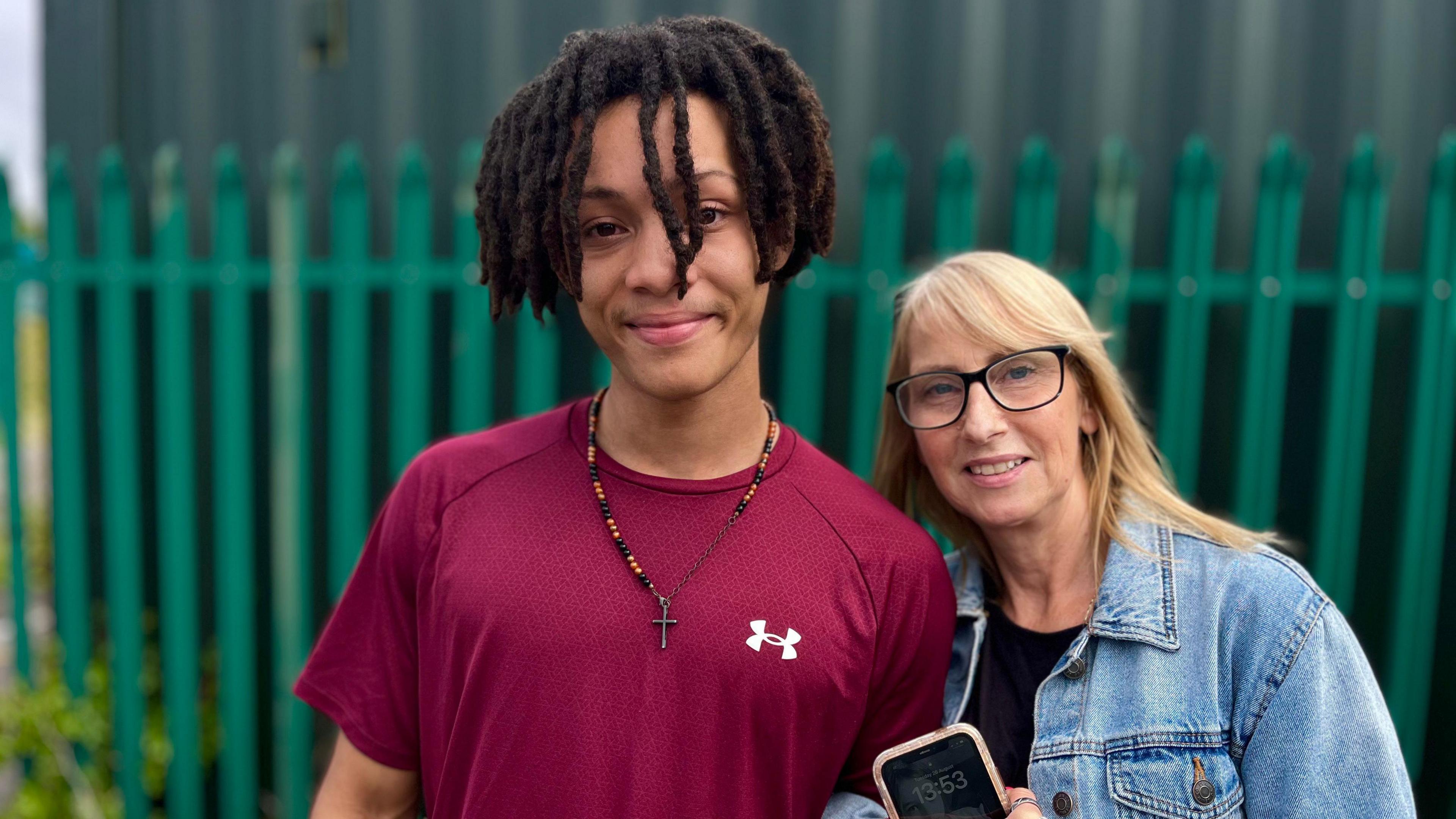 A boy in a purple top with a crucifix on a necklace, next to an older blonde woman in a denim jacket. Their arms are linked.