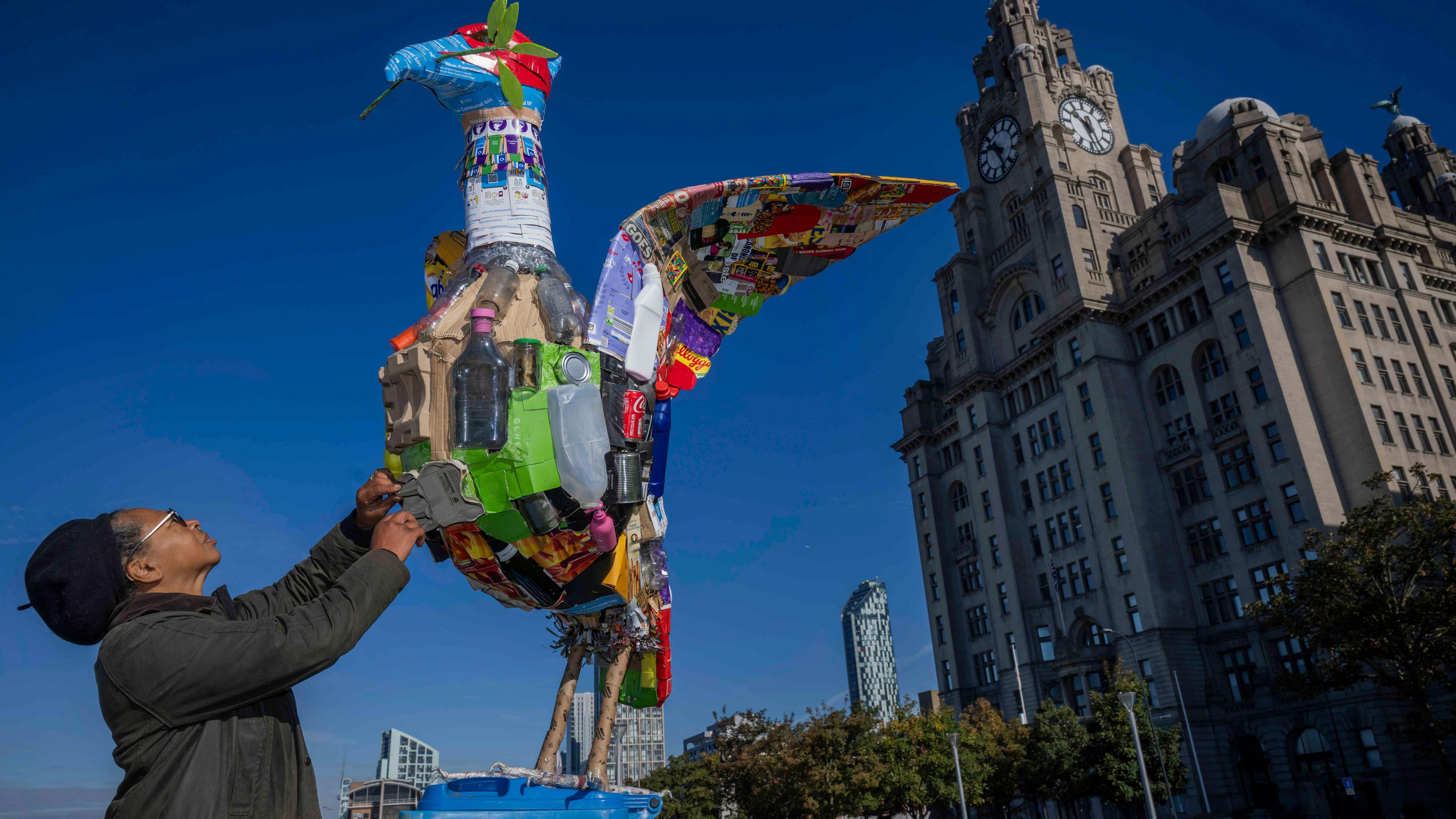 Faith Bebbington reaches towards a multi-coloured sculpture of a Liver Bird made from plastic bottles, cardboard and boxes. The Liver building is in the backdrop on a sunny day with bright blue skies.