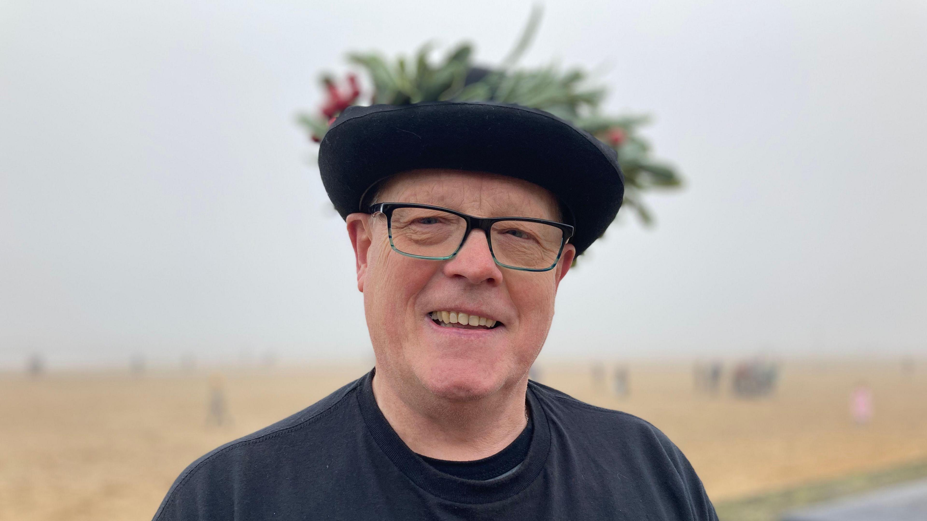 A man in a black t-shirt wearing black glasses and a hat with foliage on. He is smiling at the camera. There is a beach in the background.