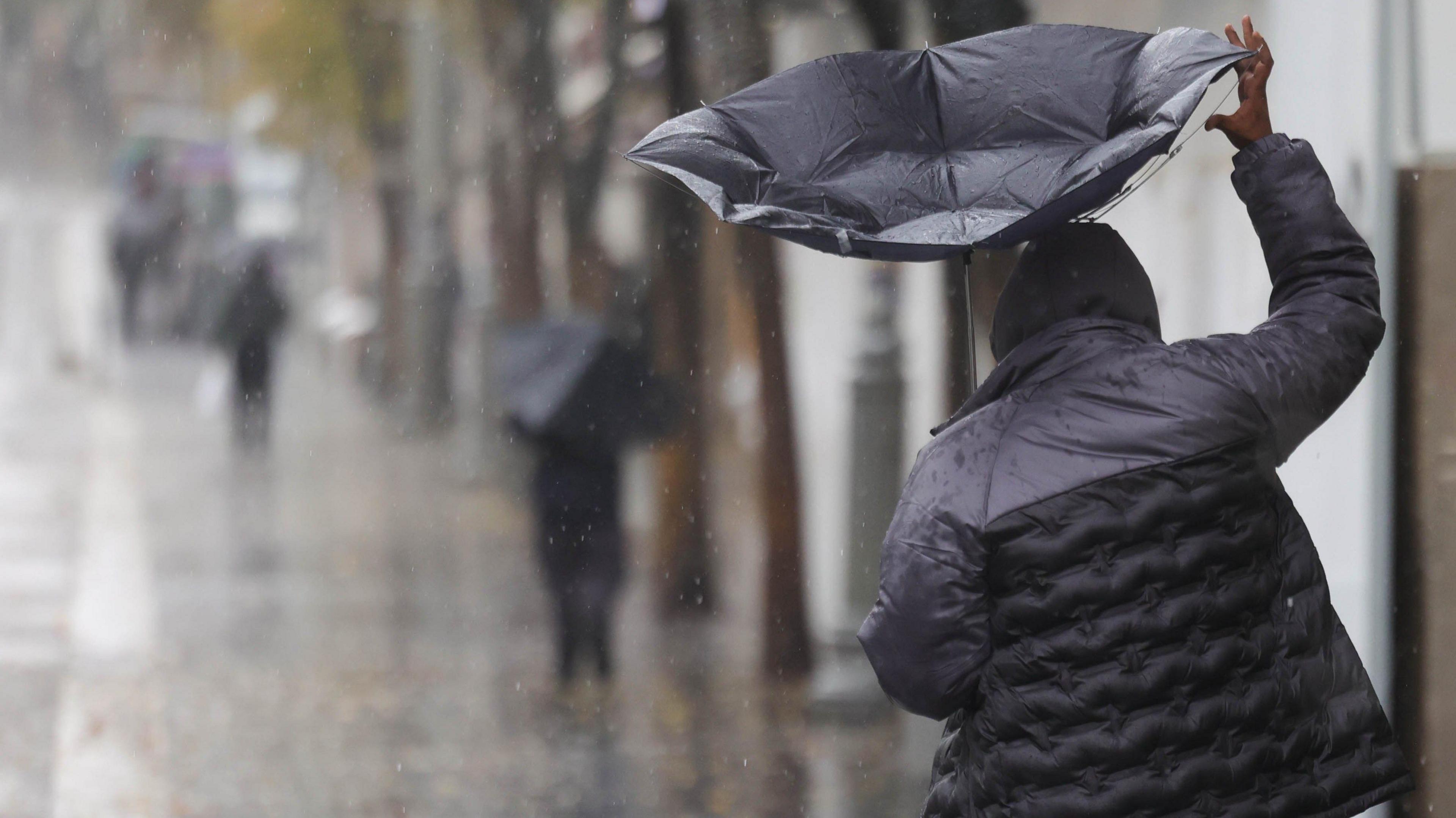 A man walks along the street in heavy rain. He holds a black umbrella which has turned inside out in the wind. Other people in the distance are also carrying umbrellas. 