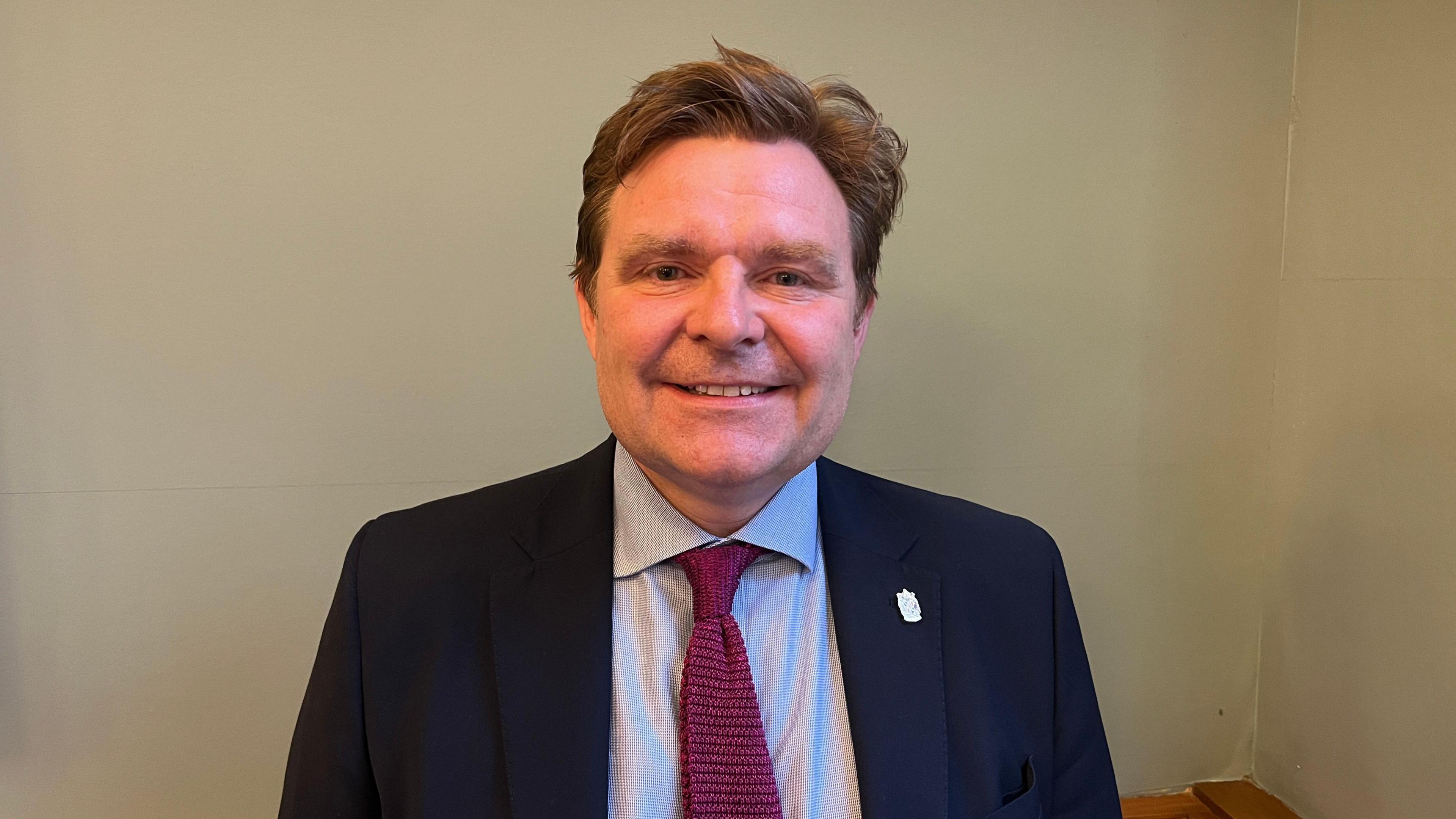 Head and shoulders shot of Councillor John Cotton, the Labour leader of Birmingham City Council. He is wearing a dark blue suit and red tie, standing against a plain cream background.