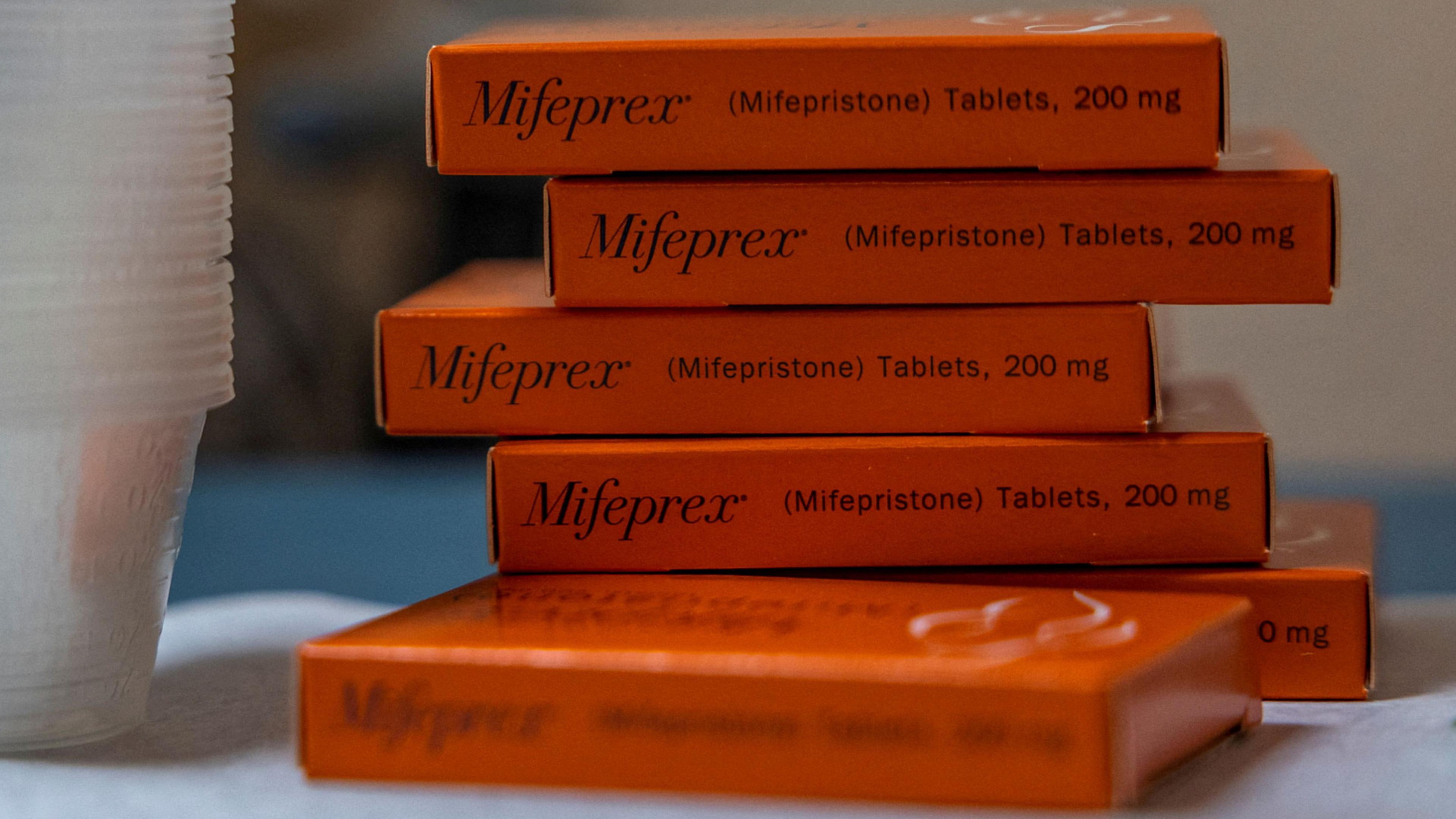 A stack of orange tablet boxes on a table with Mifepristone labelled on them. They are next to a stack of clear plastic cups. 