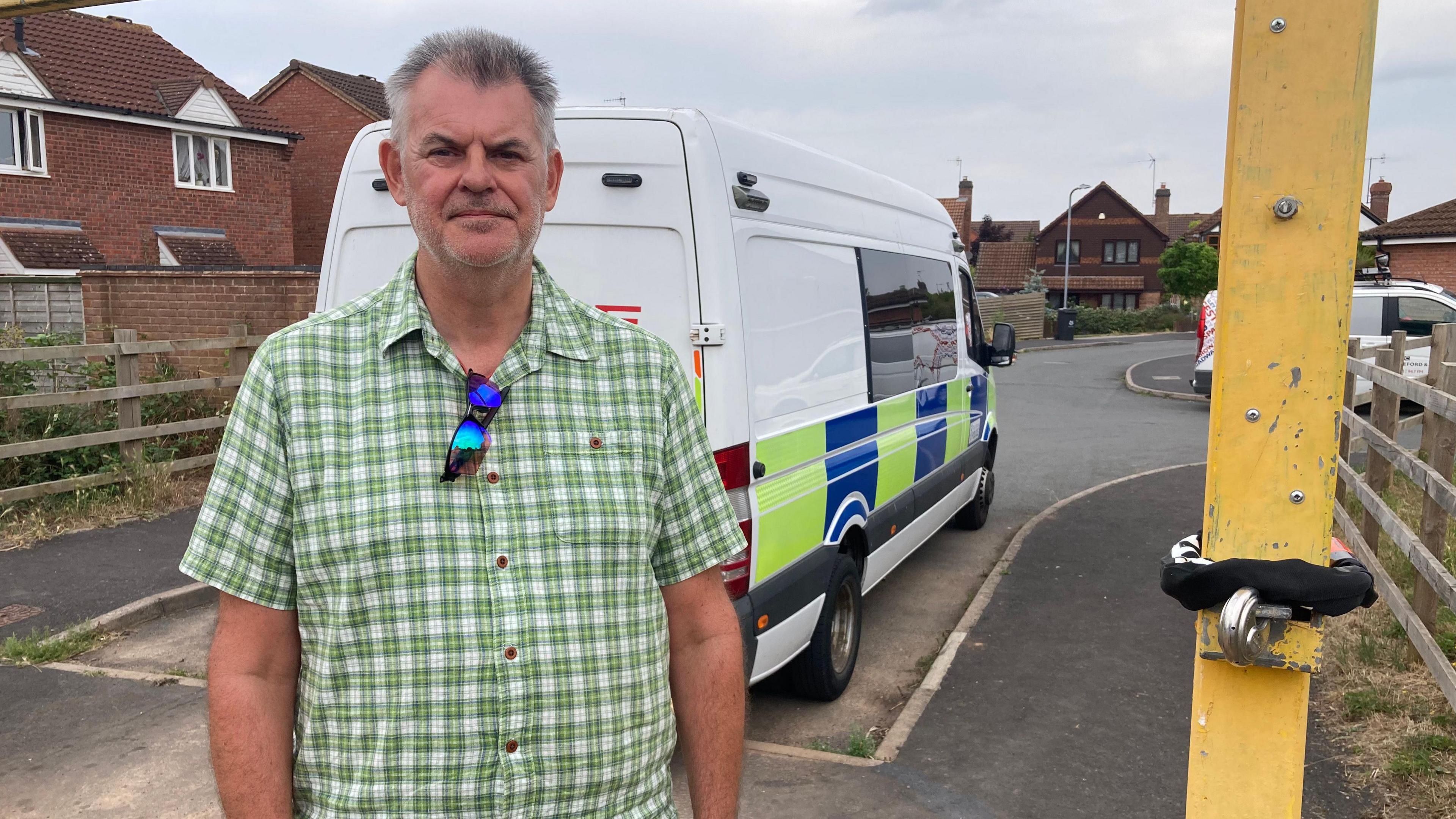 Mr Renshaw stood beneath the gate and next to the black bicycle lock that has been wrapped around the gate. A police van is parked behind him. He is wearing a green and white checked shirt with blue tinted sunglasses hanging from his shirt. 