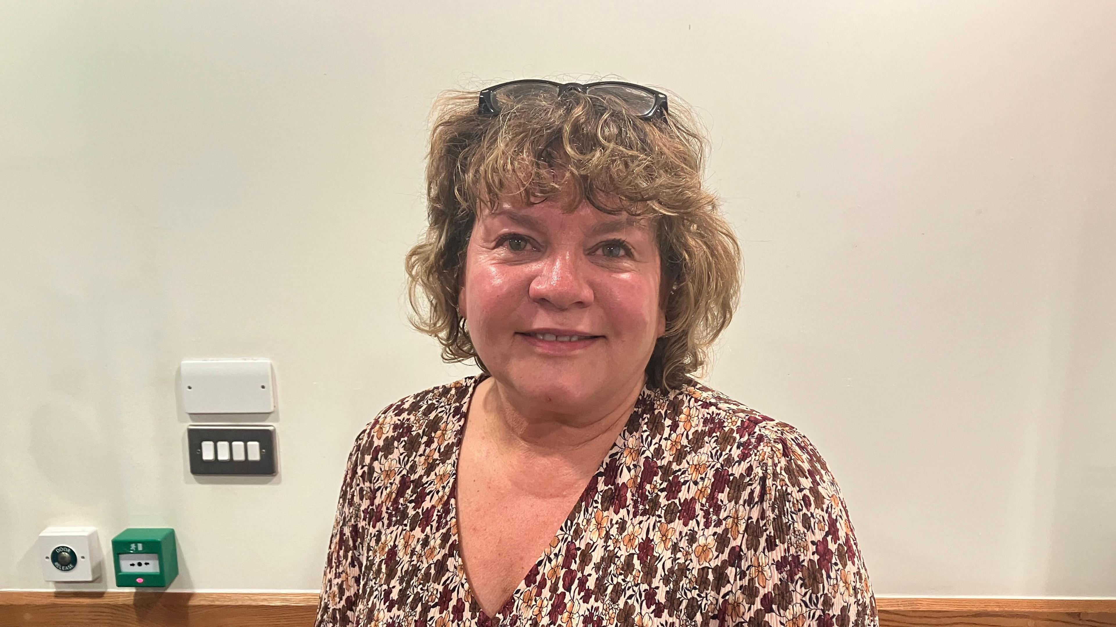 A picture of a woman with short curly hair and her glasses on her head. She has a blouse on with brown and yellow flowers on it.