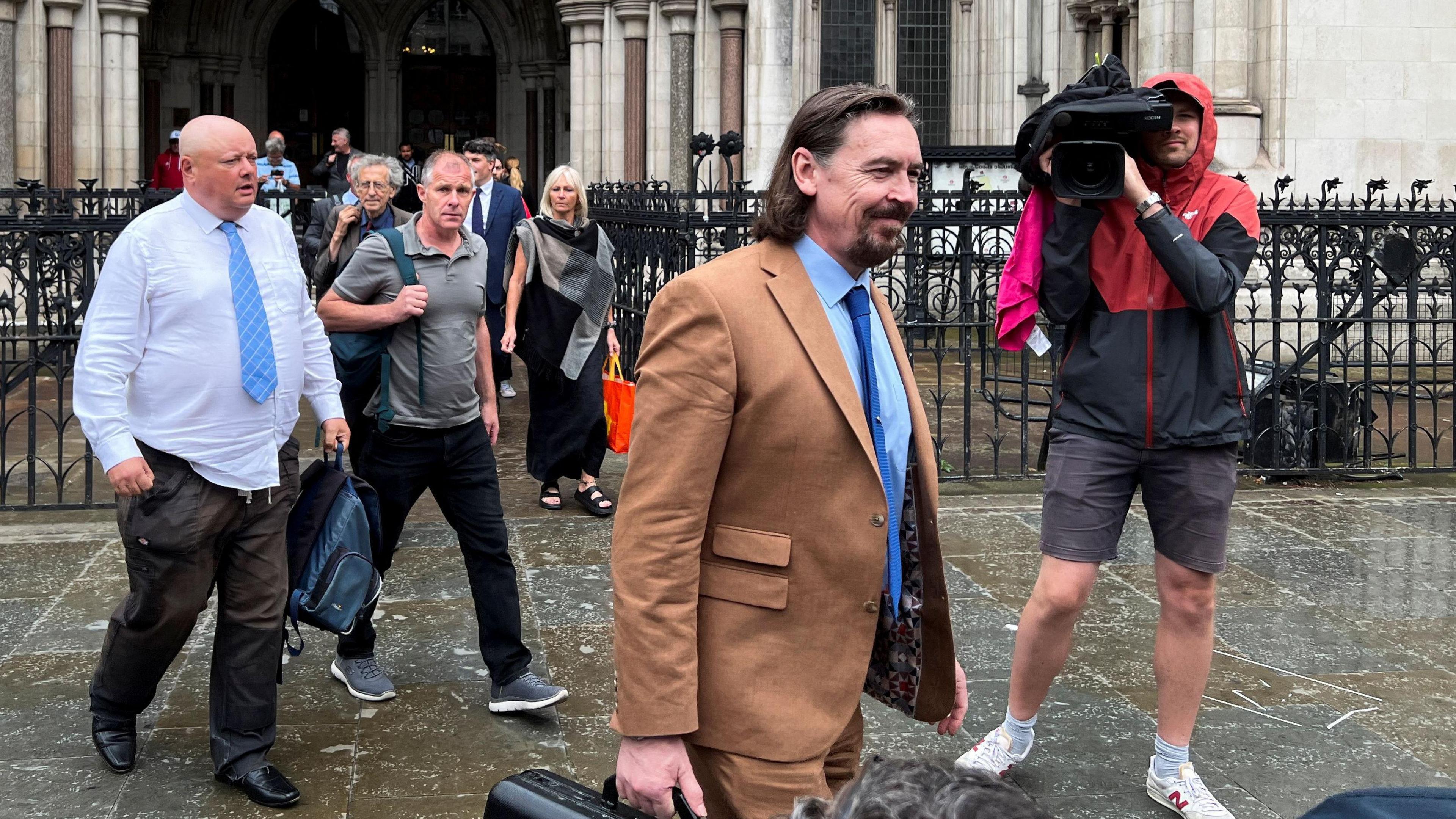 Richard Hall smiling to a cameraman, carrying a briefcase outside court