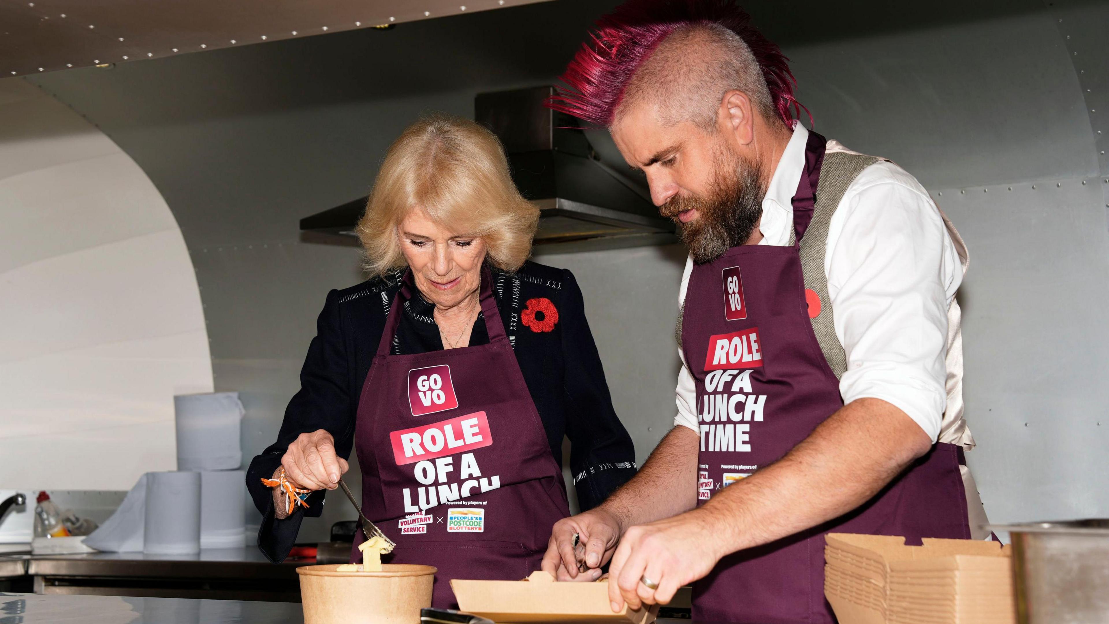 Queen Camilla, wearing a navy blue dress and a poppy with a burgundy apron, is seen talking to Ben Newman who has a pink mohawk and wearing a white shirt and grey waistcoat plus a burgundy apron. They are standing outdoors, with other people visible in the background.