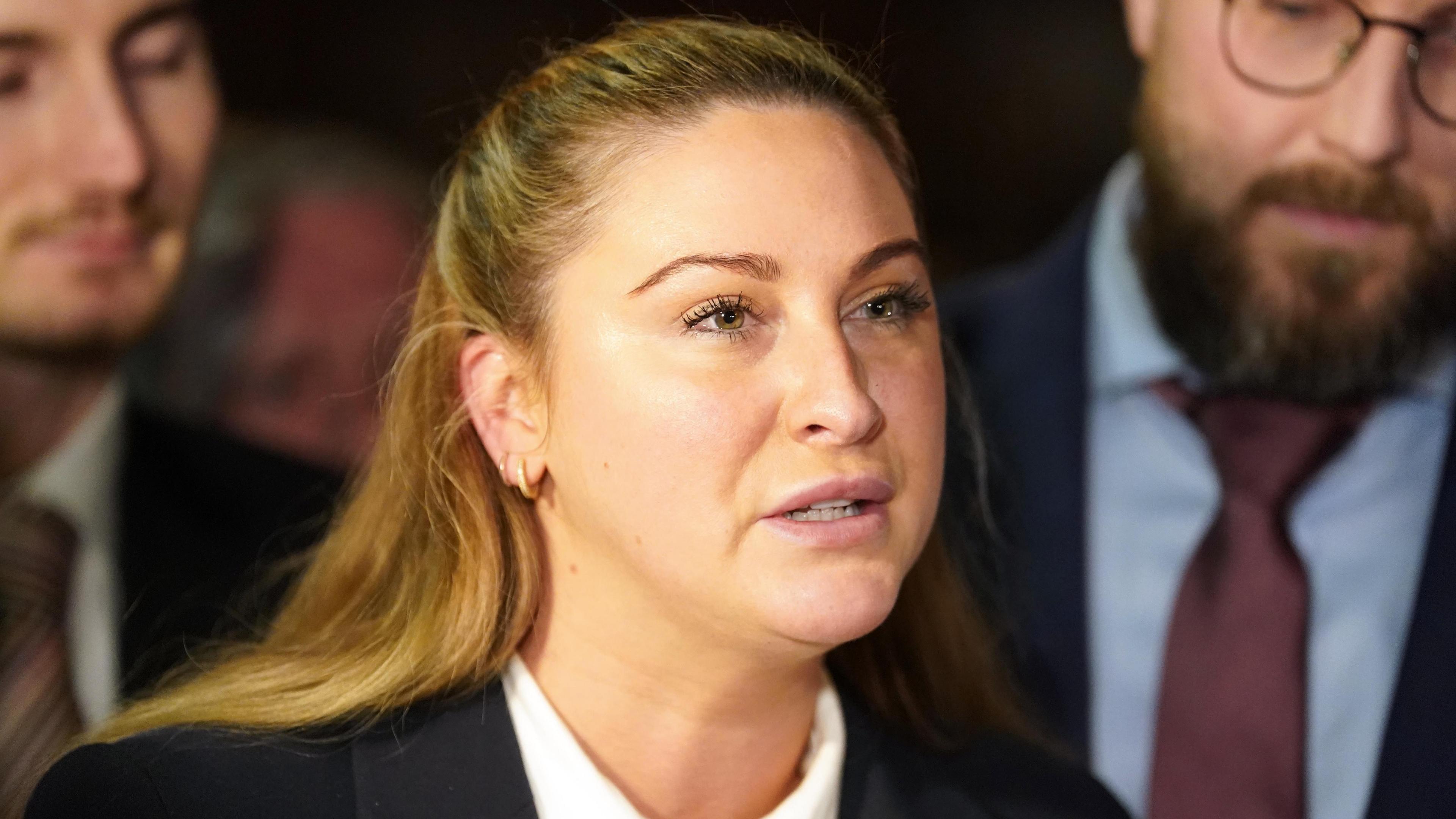 A woman outside court, she is wearing a white shirt and black blazer. She has blonde hair