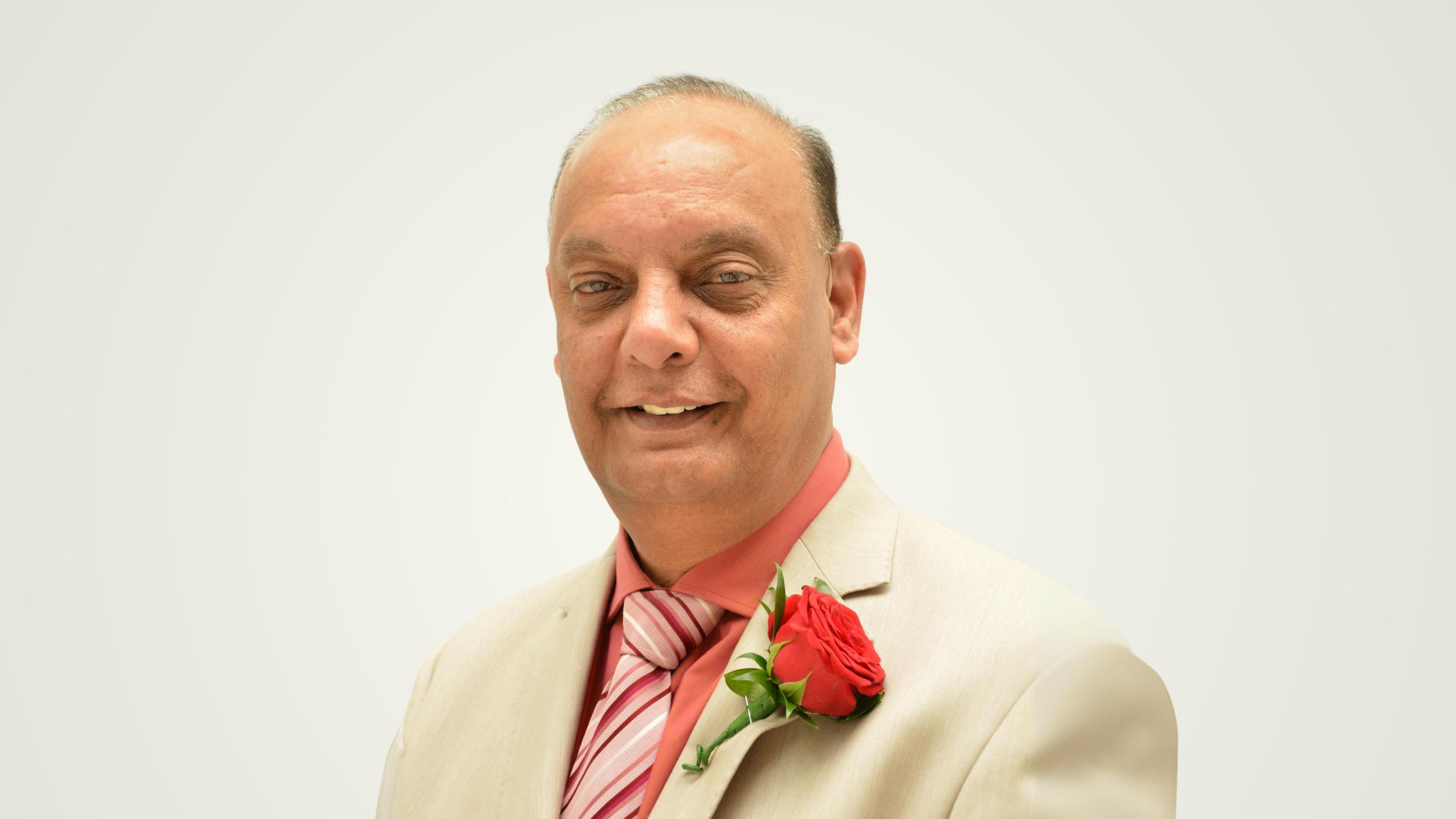 Shiraz Khan wears a cream suit blazer with a coral/orange shirt and similar colour tie. He wears a large red rose pinned to his blazer.