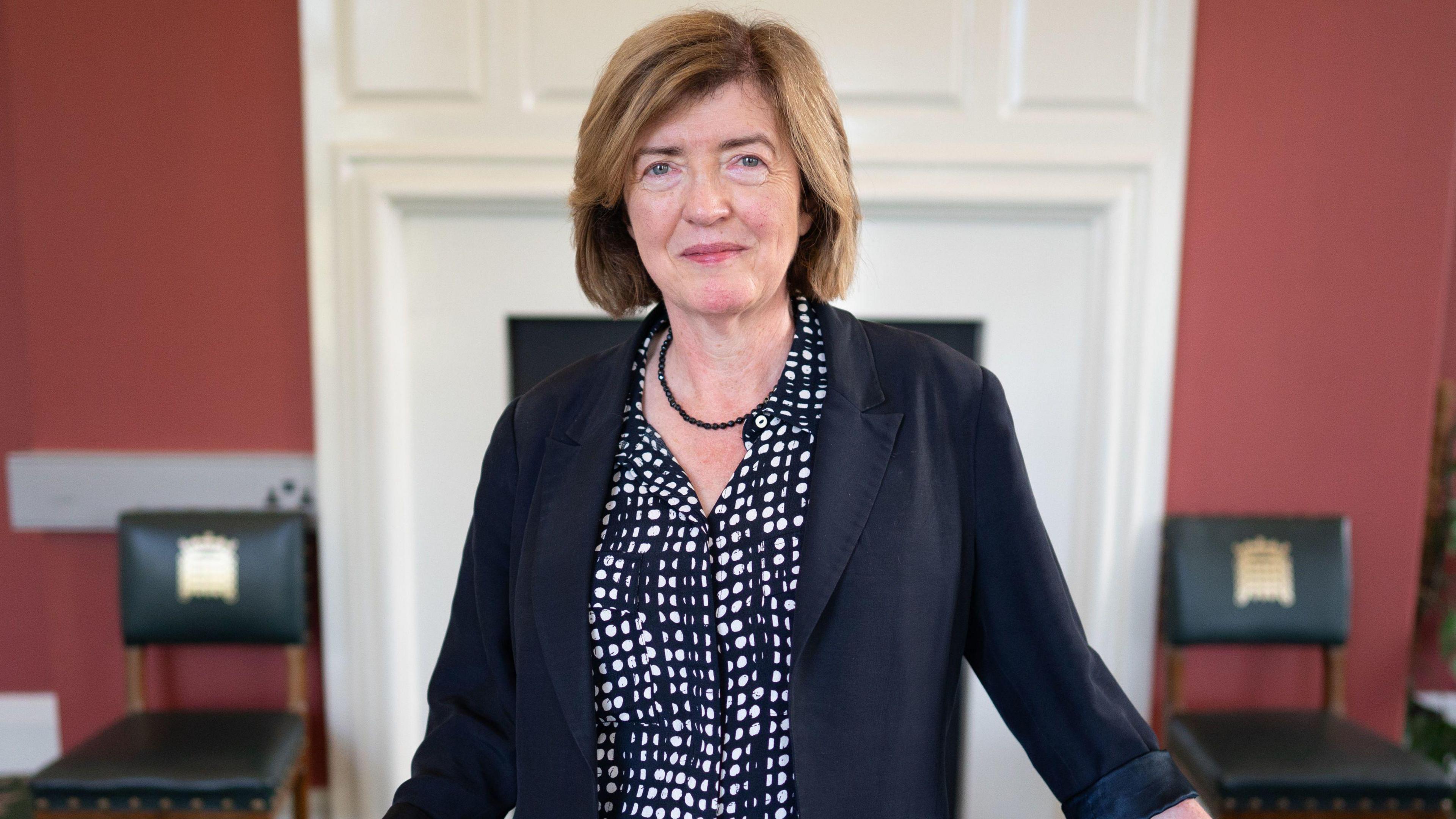 Sue Gray standings in a red room smiling at the camera. She has short, light brown hair and is wearing a black beaded necklace, a black suit jacket and a black and white polka dot shirt.