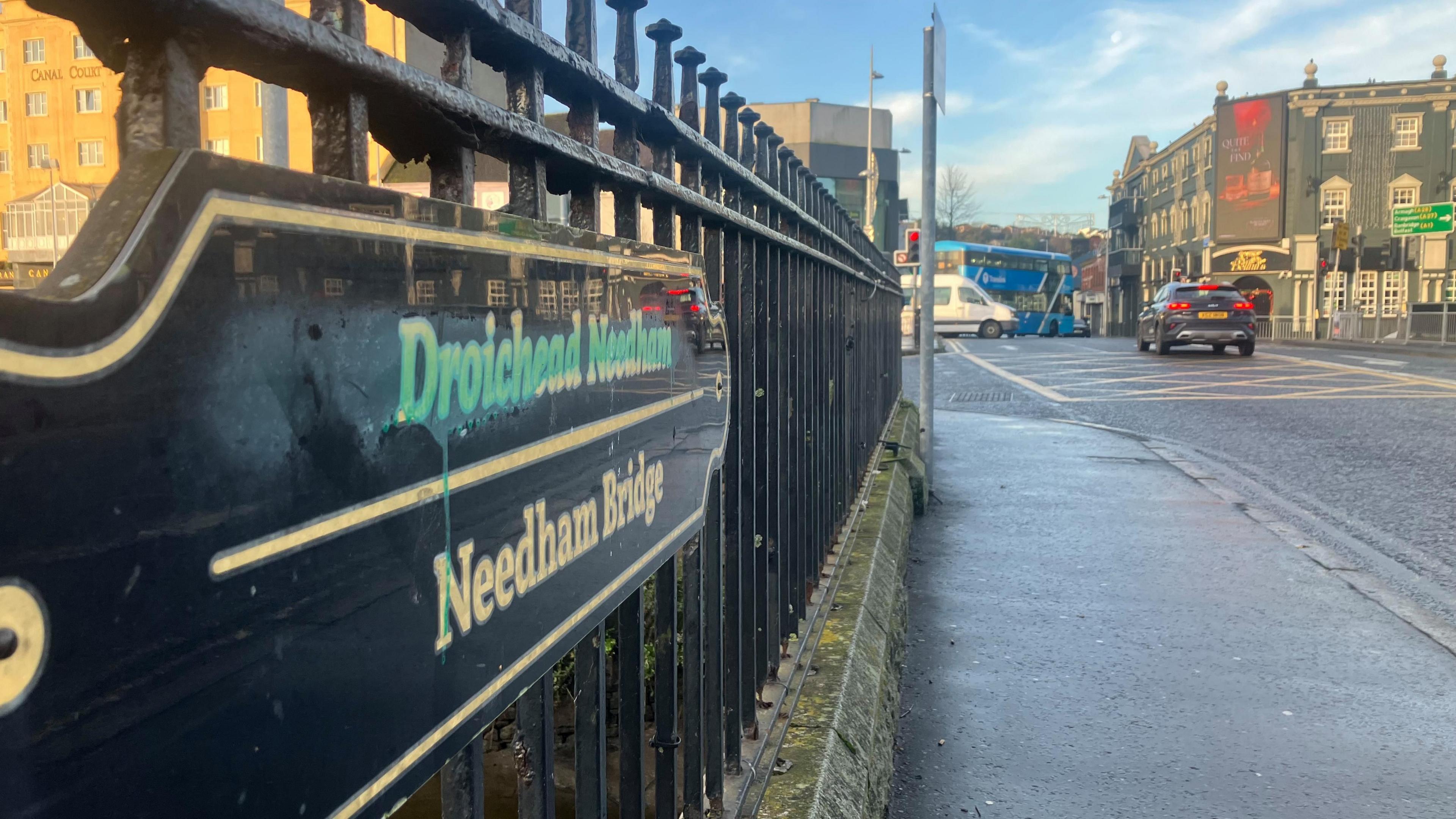 A black gate with a sign which reads Needham Bridge, road and traffic in the background 