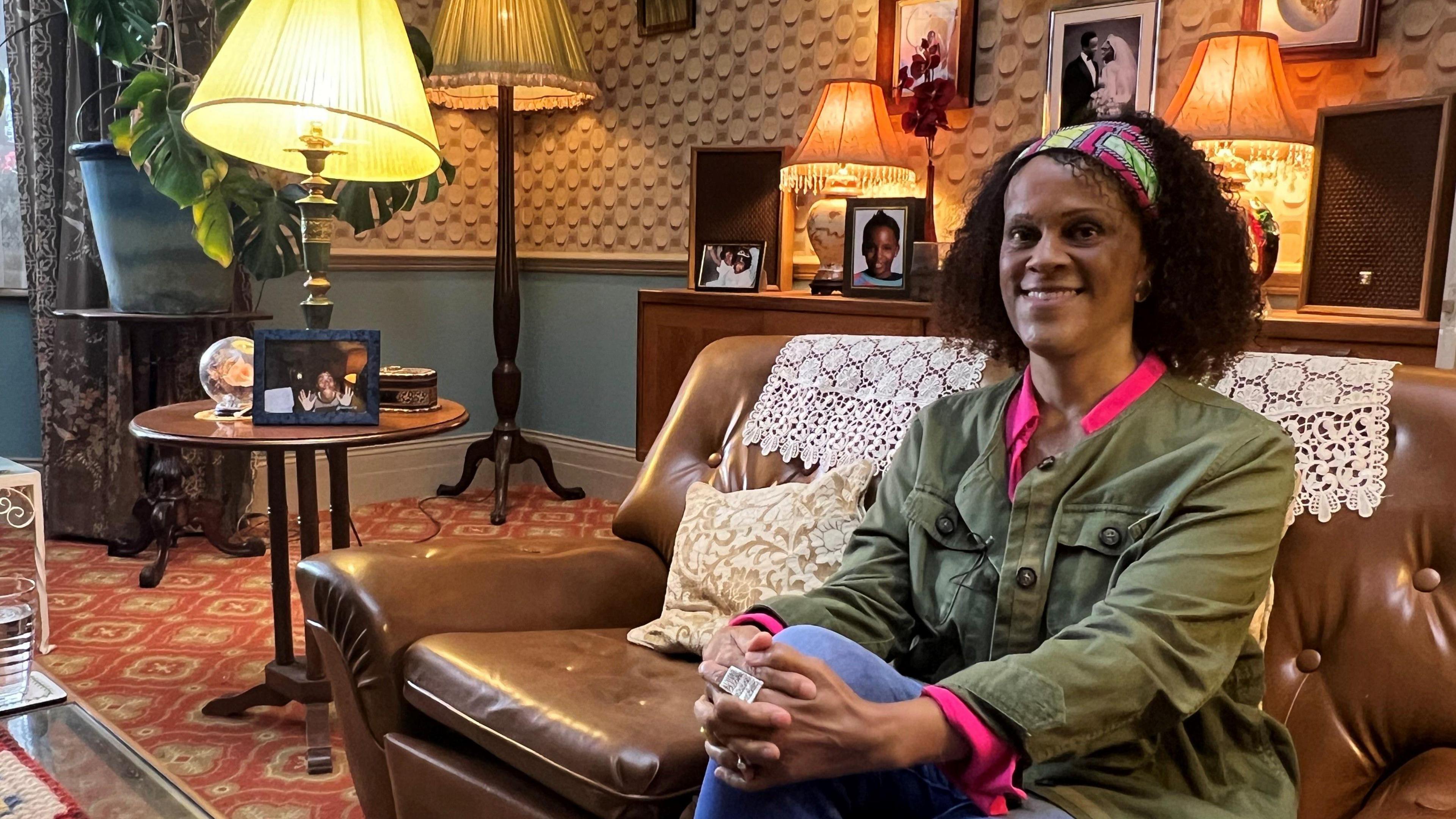 Bernardine Evaristo sitting on a brown leather sofa wearing a green jacket, blue jeans and a pink shirt in a living room