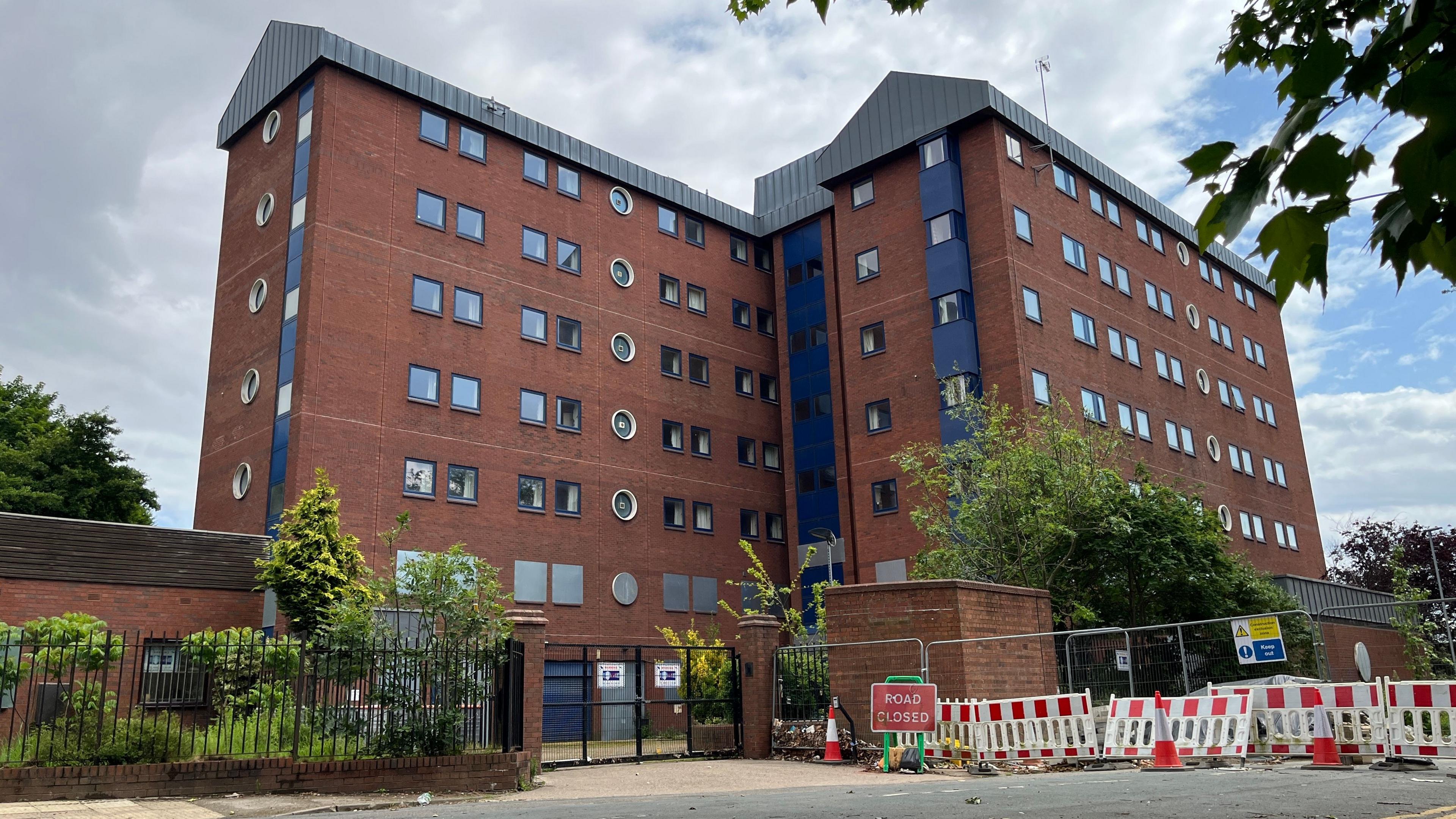 Two large tower blocks made of red bricks, blue cladding and grey panels stand behind a metal fence. The lower doors and windows are covered with metal sheeting. A sign next to the gate reads 'Road closed'.