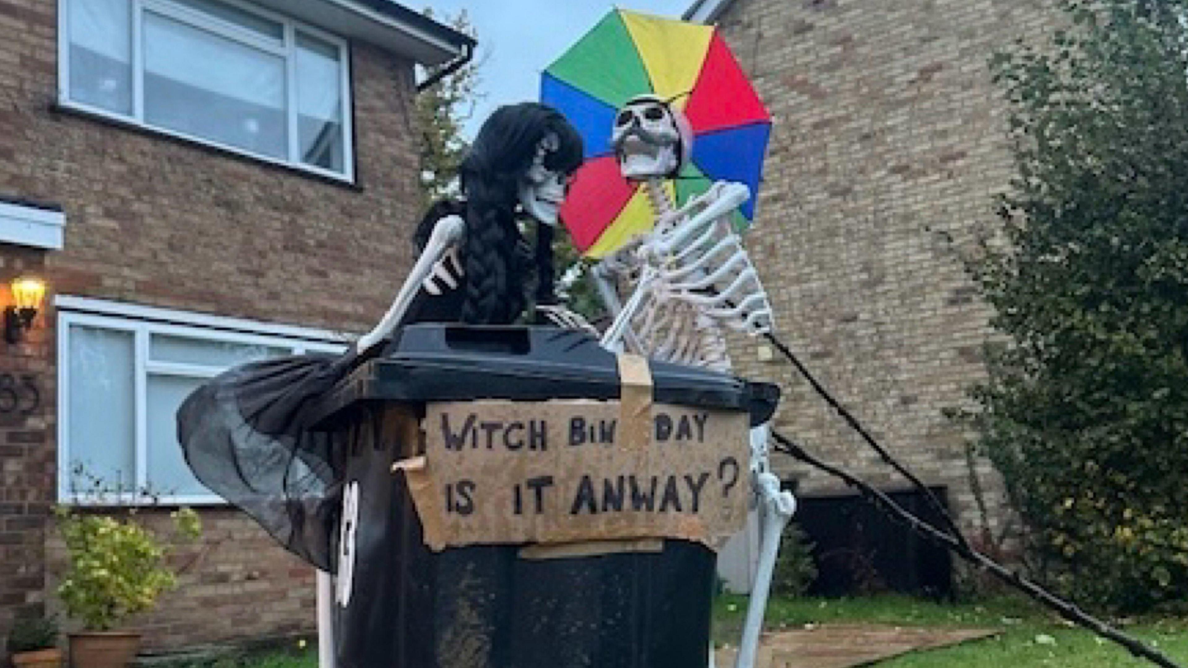 Two skeletons stood by a bin, one is holding an umbrella. There is a sign that says 'Witch bin day is it anyway?'