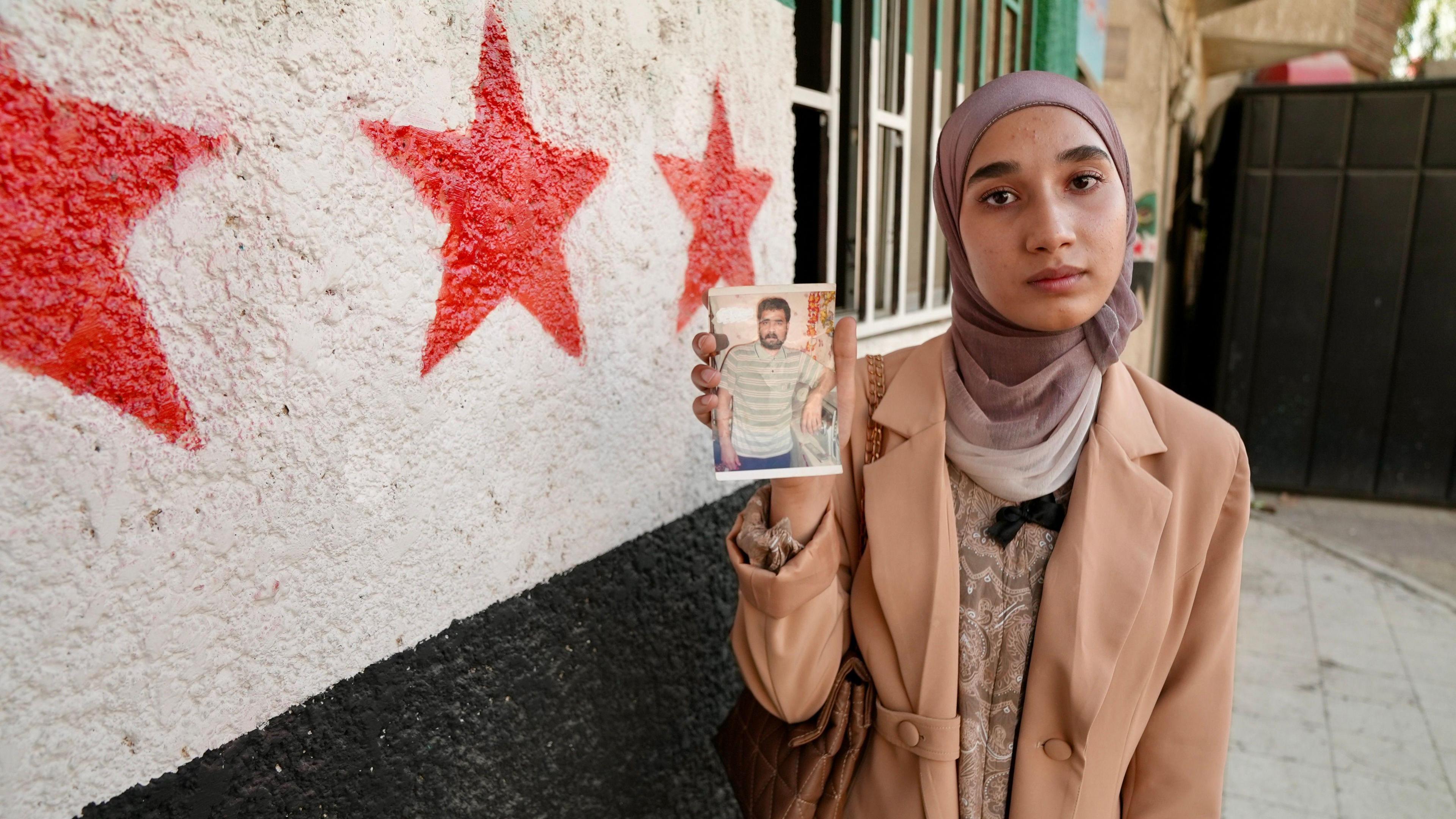 A young woman holds up a photo of a missing man. She is standing against a wall with stars from the Syrian flag painted on it.