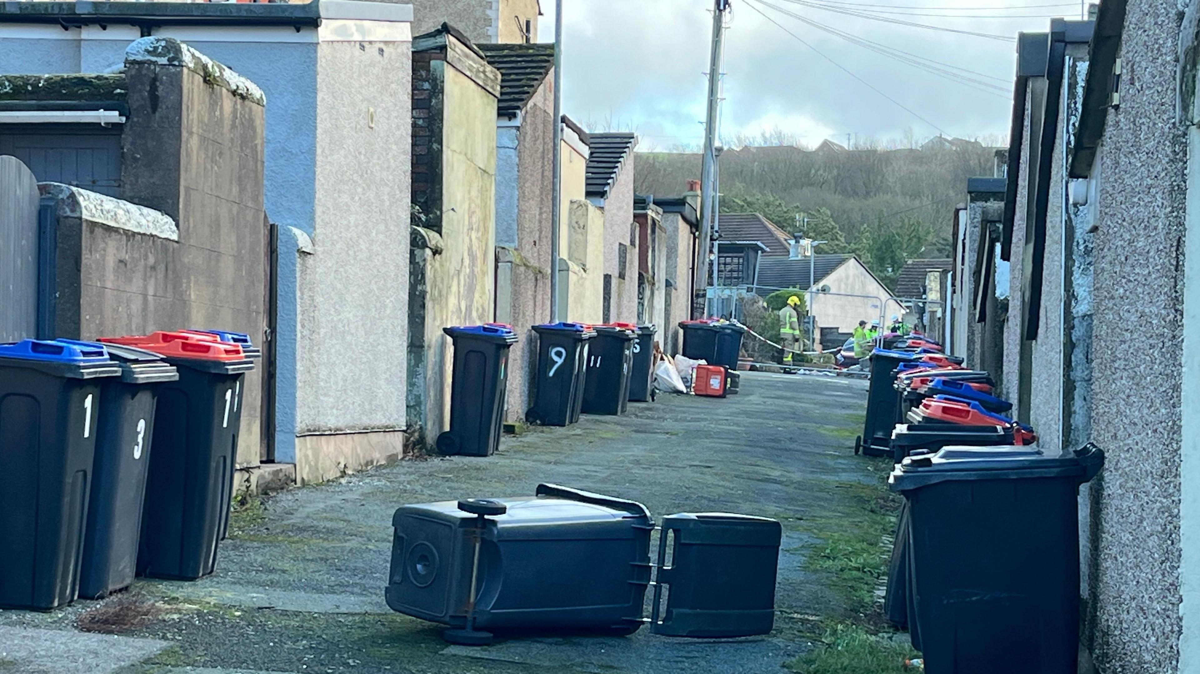 A back lane with wheelie bins, some on their side. At the bottom of the lane there is a fence and firefighters wearing high-viz and helmets are working behind it.