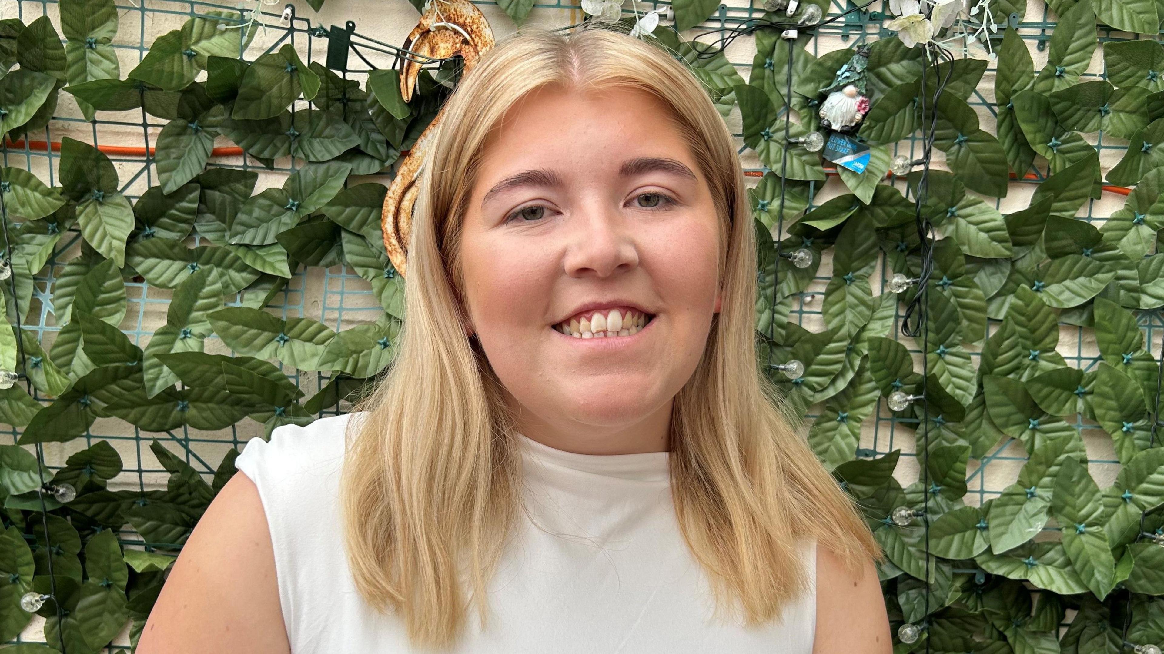 A girl in a white top smiling at the camera. The background is of green leaves. It is a head and shoulders shot of her.