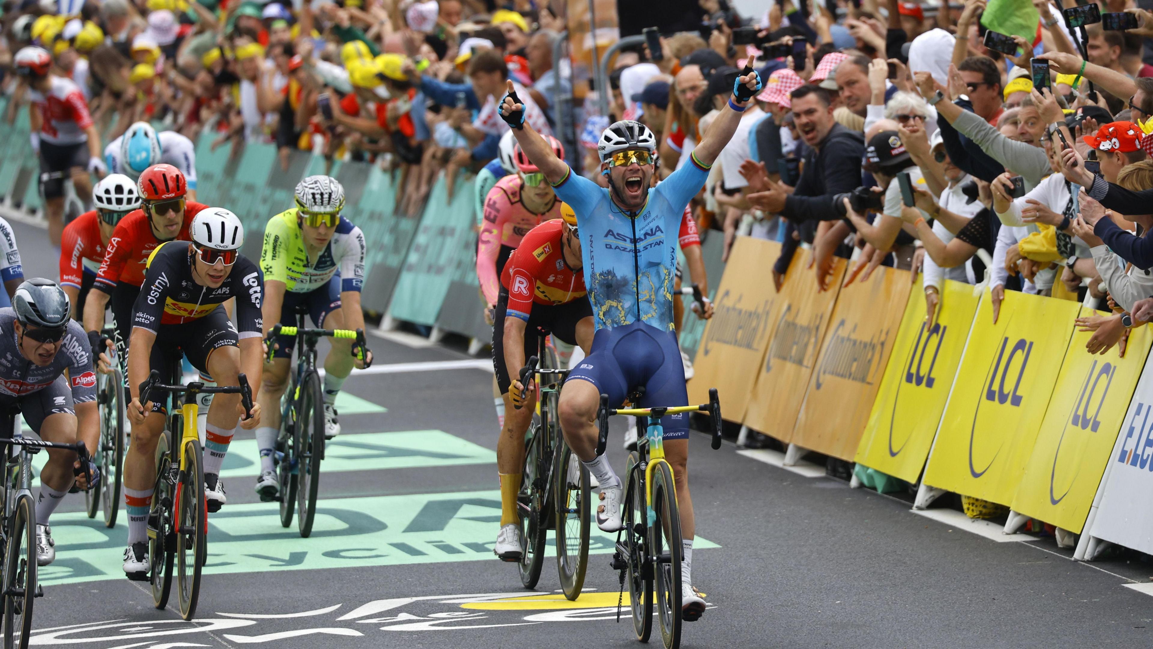Sir Mark Cavenidsh, wearing a blue cycling top and shorts and a white helmet, holds his arms up and smiles as he wins a Tour de France stage ahead of a number of other cyclists and in front of a cheering crowd