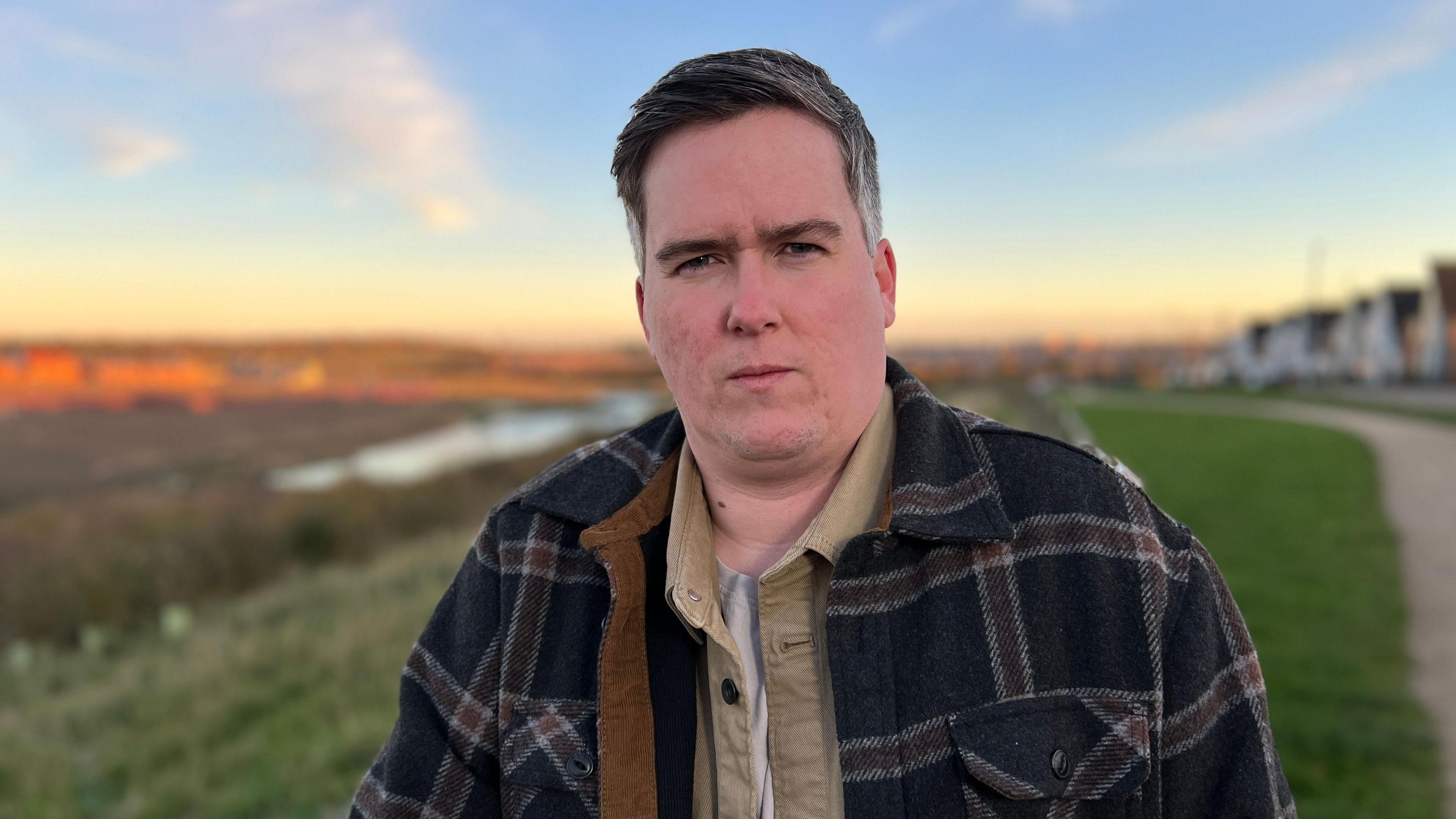 A man in a beige shirt, and navy tartan coat, stands on a strip of grass, looking straight at the camera.