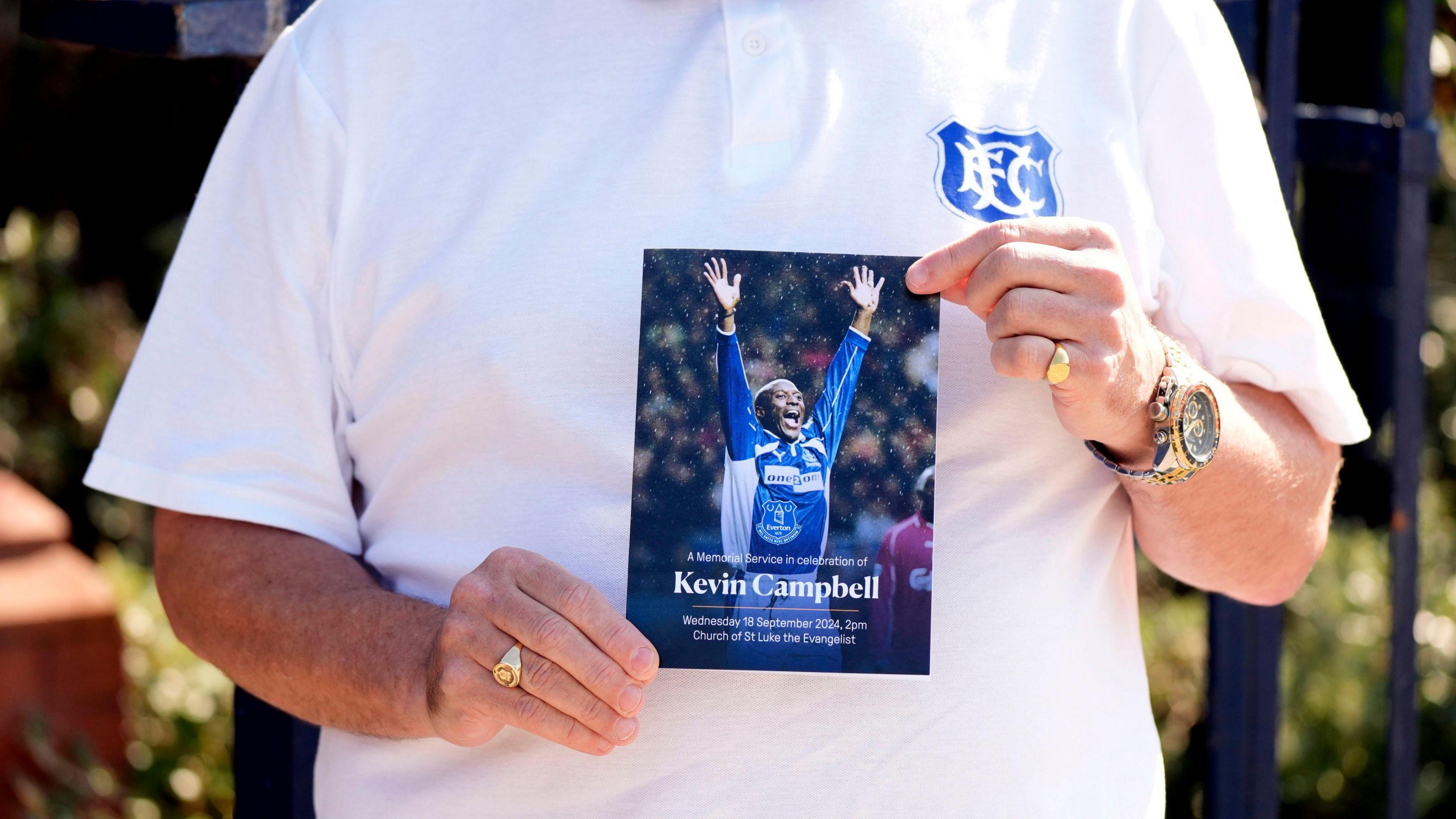 A man in a white polo-neck t-shirt with an Everton logo holds a copy of an order of service, which feature of a photo of Kevin Campbell in an Everton kit during his playing days raising both arms in the air in celebration. 