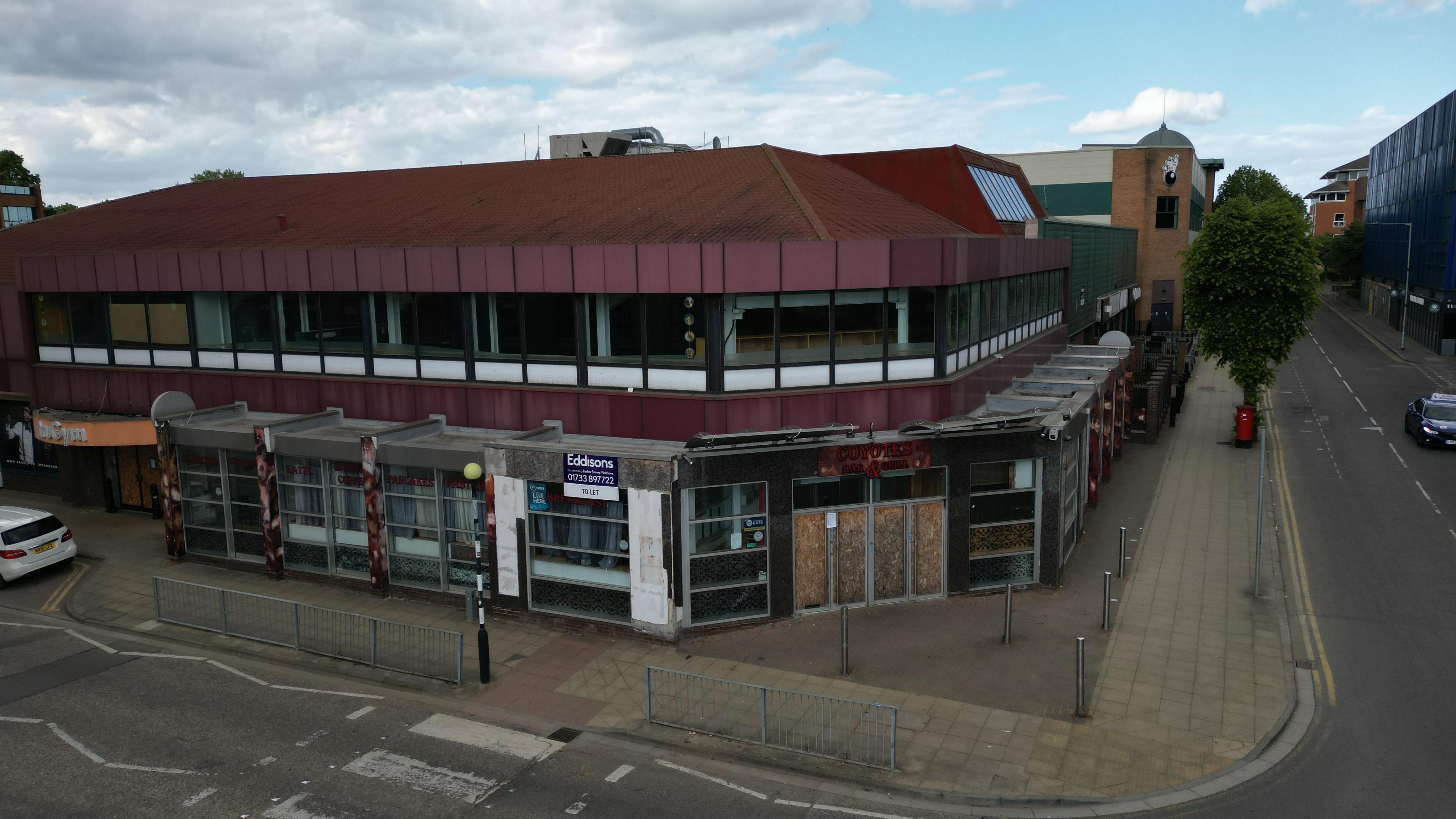 A large derelict building stands on a corner plot. It has a boarded-up door and bars over the windows.
