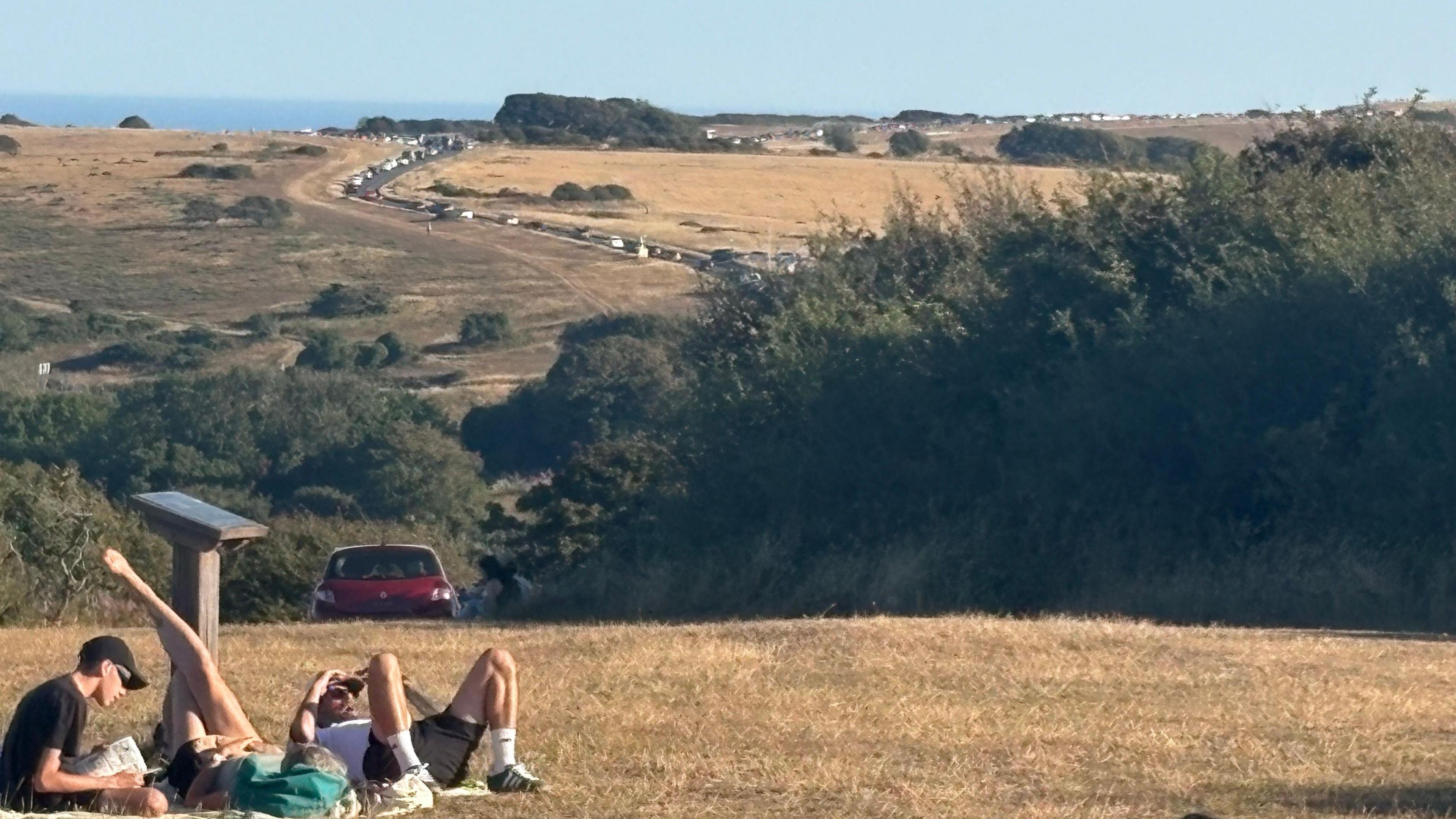 Several people sit and lie on dry grass on high ground near Beachy Head in East Sussex. A long queue of cars stretches along a road beyond them, with the sea visible in the distance.