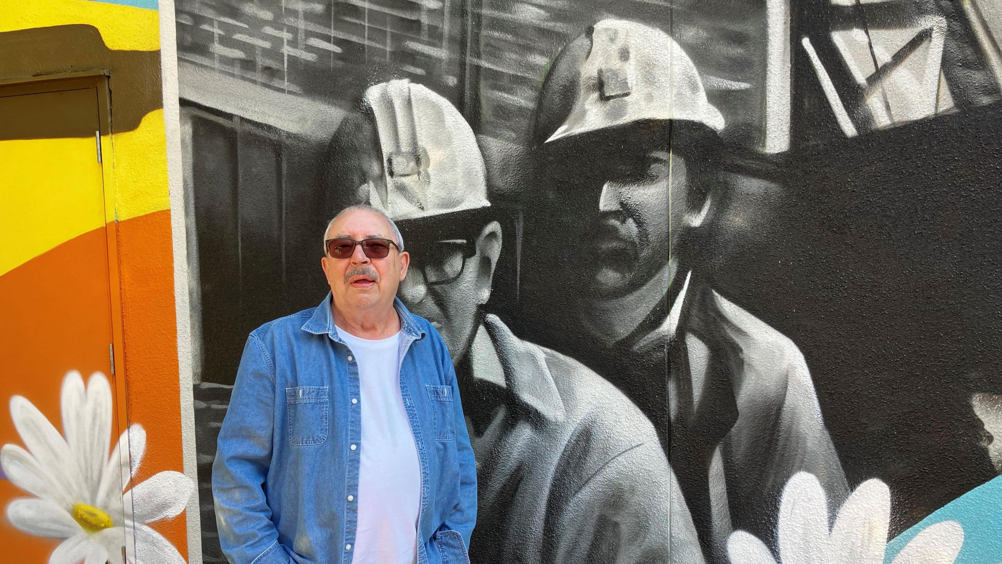 Malcolm Sword, a man wearing sunglasses and a denim jacket over a white T-shirt is stood against a black and white mural of two miners wearing helmets, which also has a brightly-coloured section to the left hand side