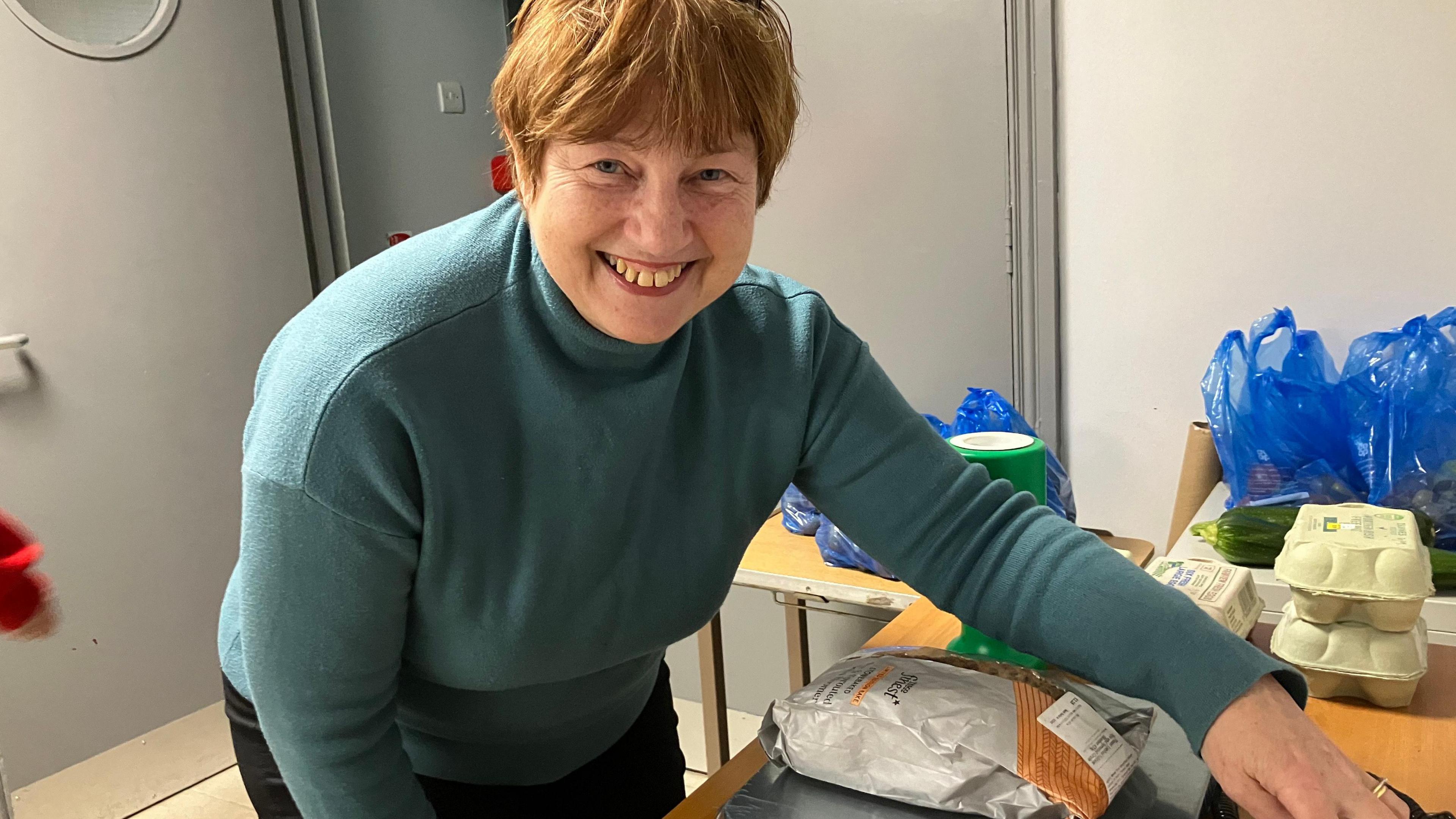 A woman is smiling straight into the camera. She's wearing a blue coloured polar neck jumper and has a pair of glasses balancing on her hair.  She is weighing a loaf of bread on a set of scales as part of the Community Fridge initiative.  Sitting beside the scales are boxes of eggs laid by the hens in the community garden.