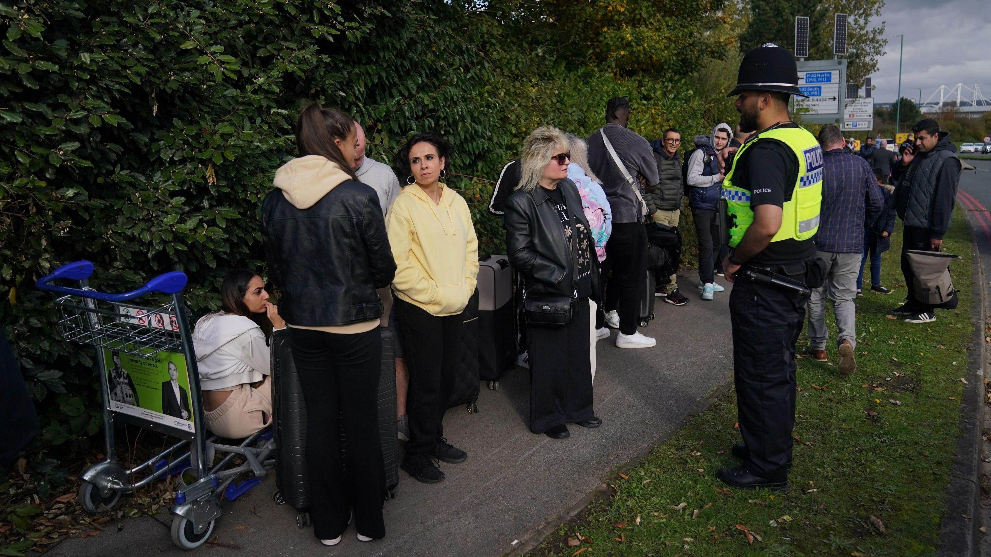 People stood on a pavement, one is sat on a luggage trolley. A police officer is stood nearby the group.