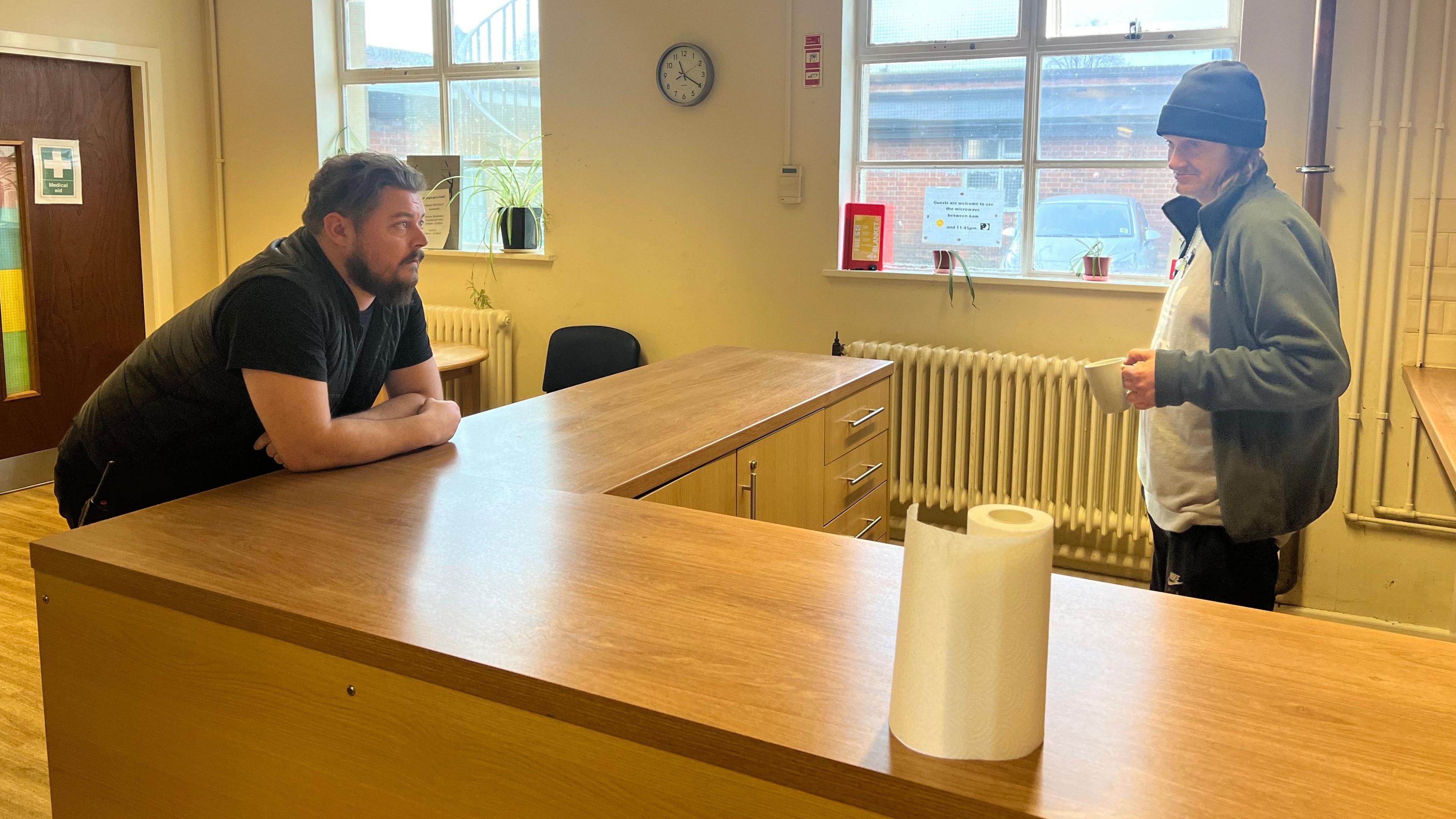 Two men speak in a kitchen area , one leans on the wooden counter, the other holds a mug behind the counter 