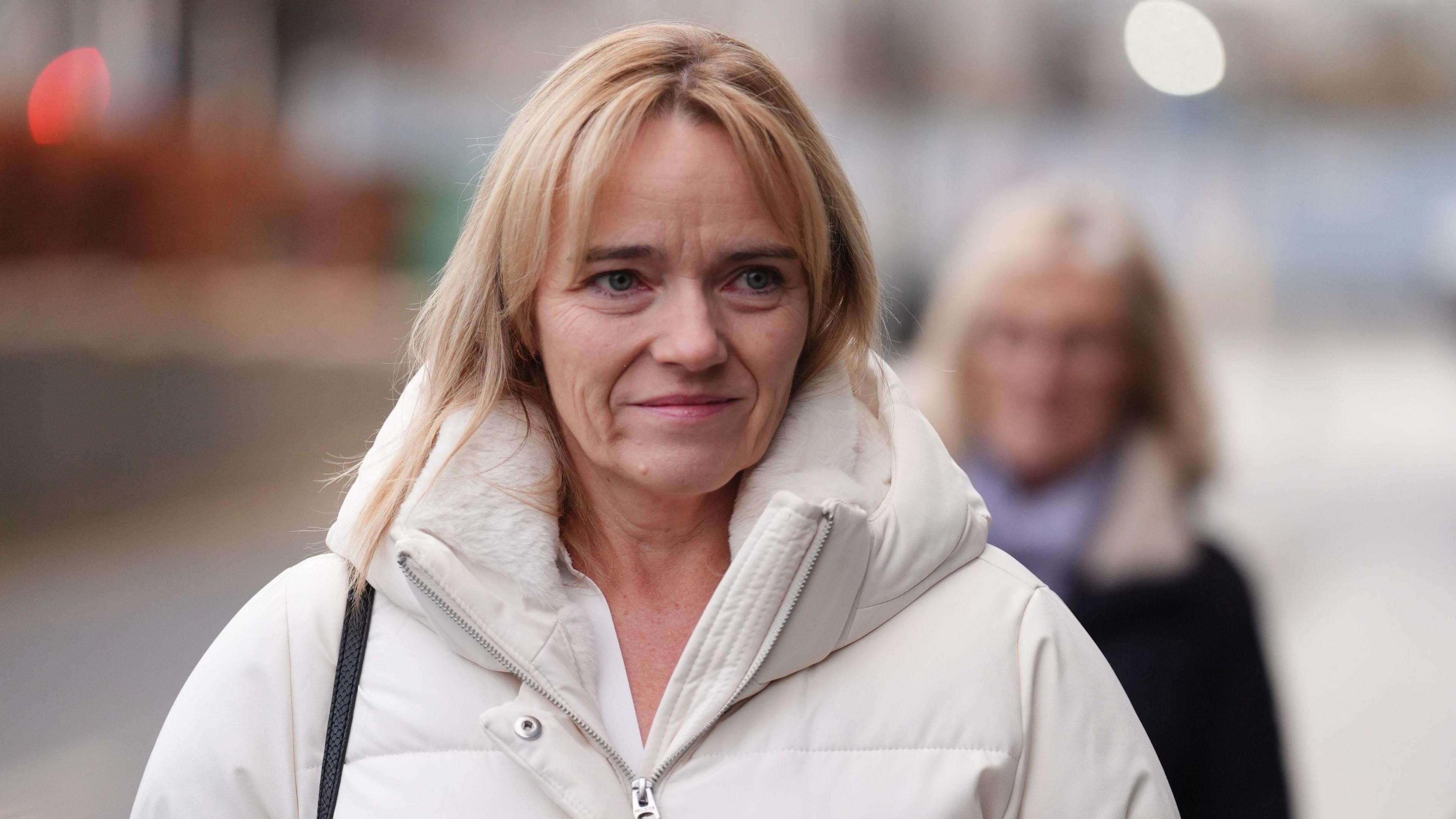 Sandie Peggie - a woman in a white jacket with blonde hair - walking down a street. She is smiling.