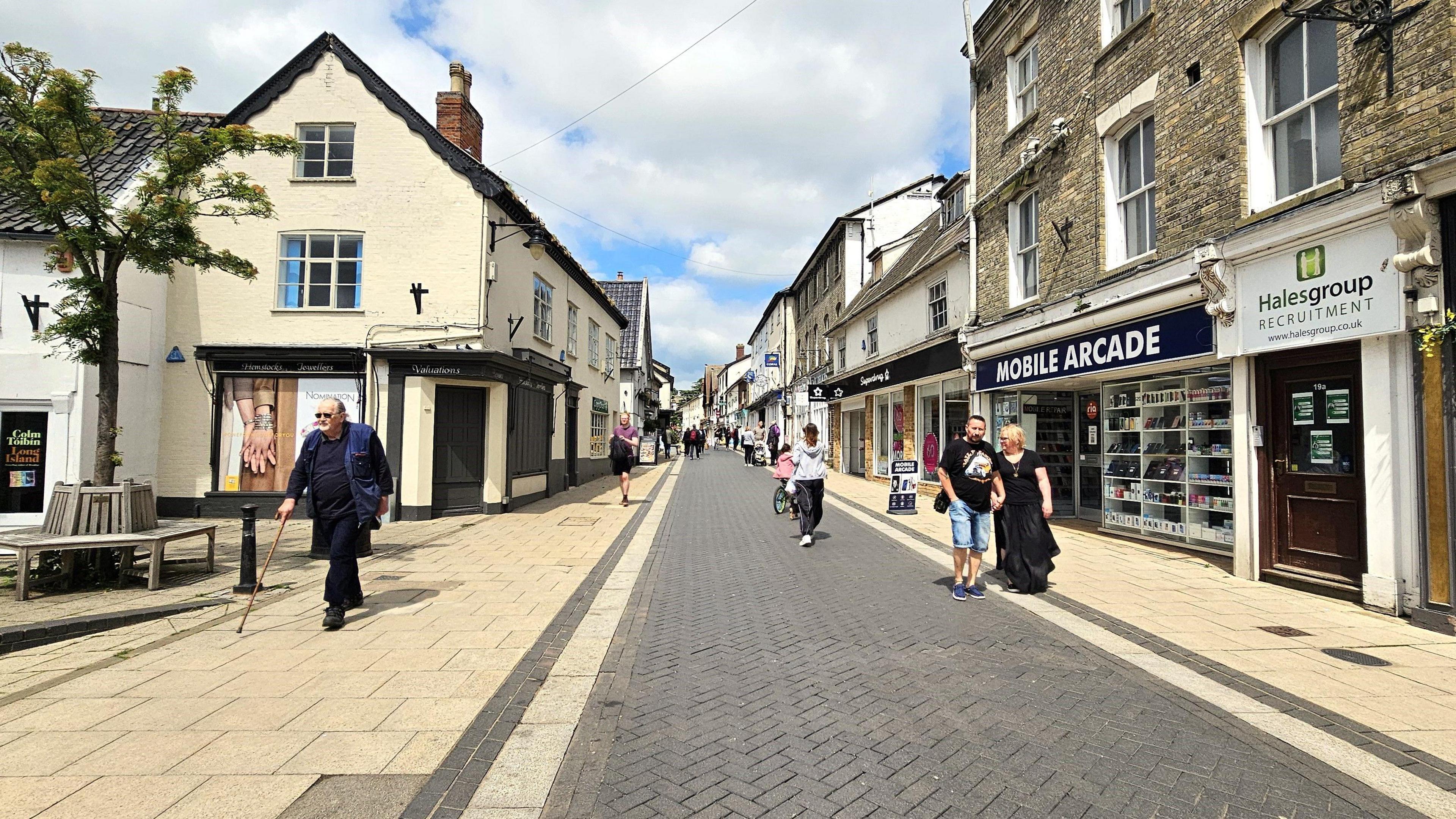 The main street in Diss, Mere Street. It's a sunny day with shops on either side of a pedestrianised area. There are a few shoppers walking in the street.