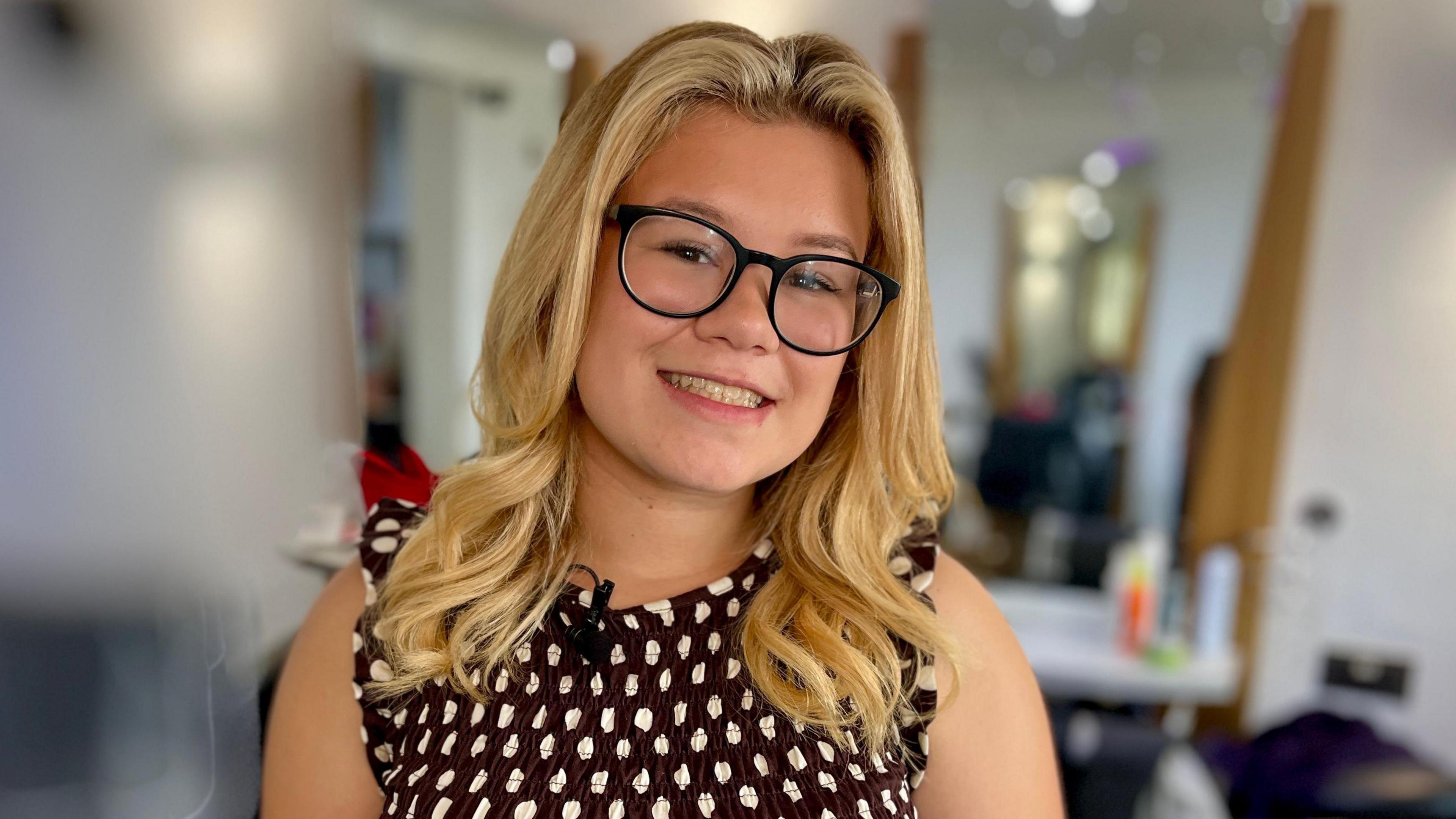 Jessica Flaxman stands in a hair salon with the background blurred out. She has long blonde hair and wears black-rimmed glasses. She is dressed in a brown dress with white polka dots.
