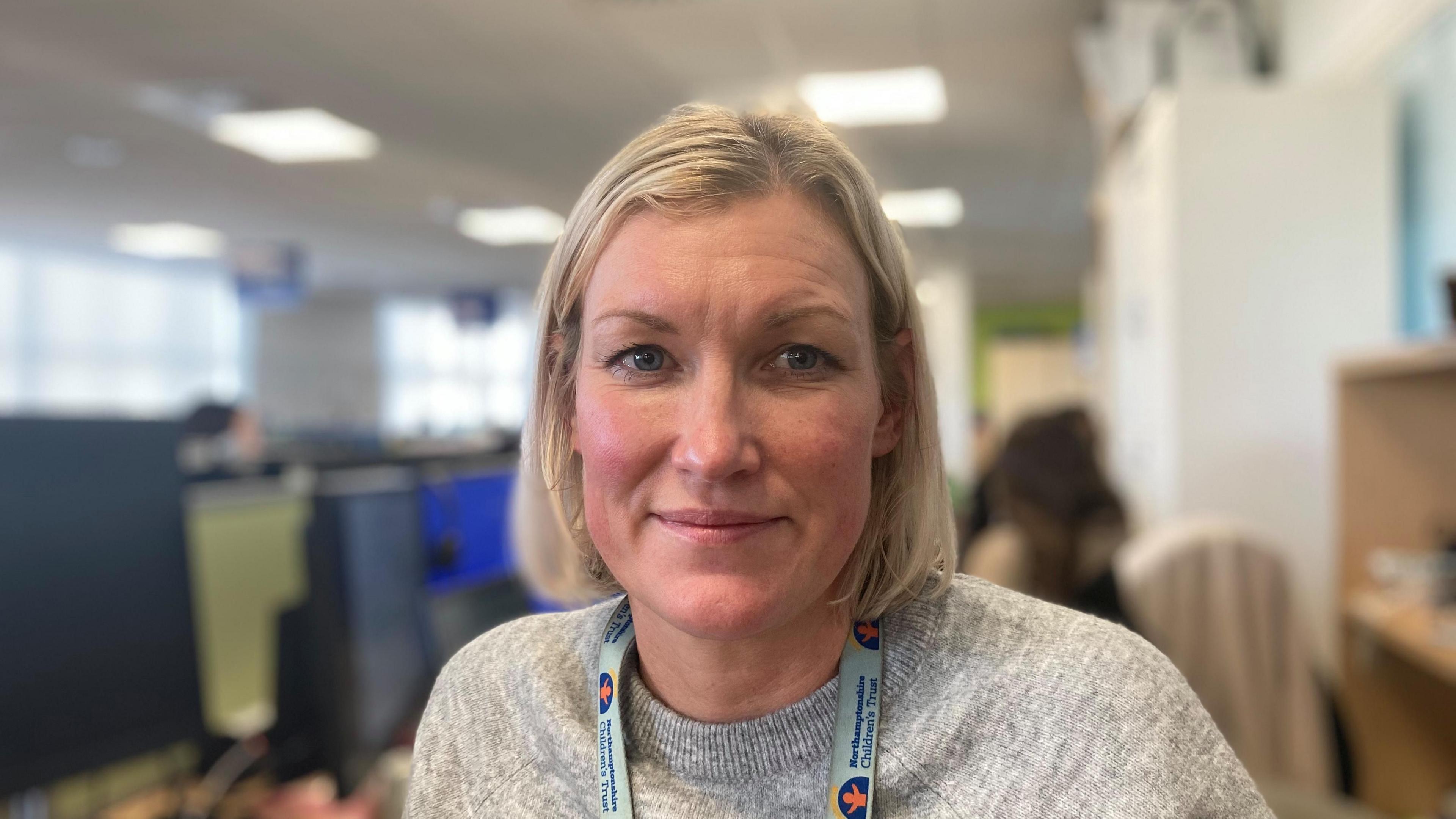 Laura Isherwood wearing a grey jumper with a light blue lanyard. People are working in the office behind her.